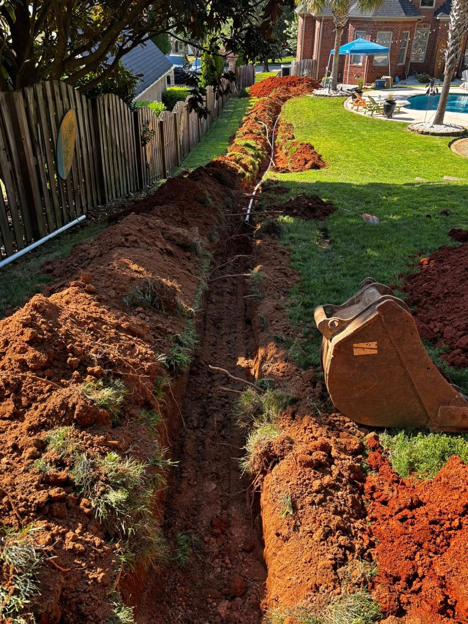 A long trench dug in a green lawn next to a wooden fence, with a small excavator bucket visible.