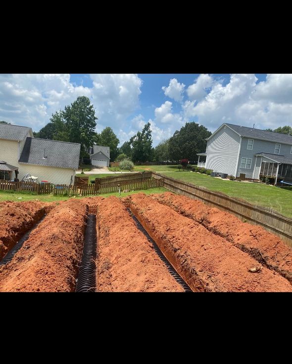 Trenches dug in red soil in a residential backyard. Black drain pipes are visible in one trench, houses and trees in the background.