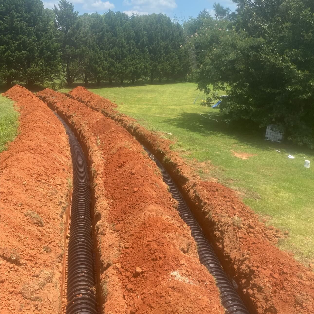 Two parallel trenches dug in red soil, with black corrugated pipes running through them, set in a grassy yard.