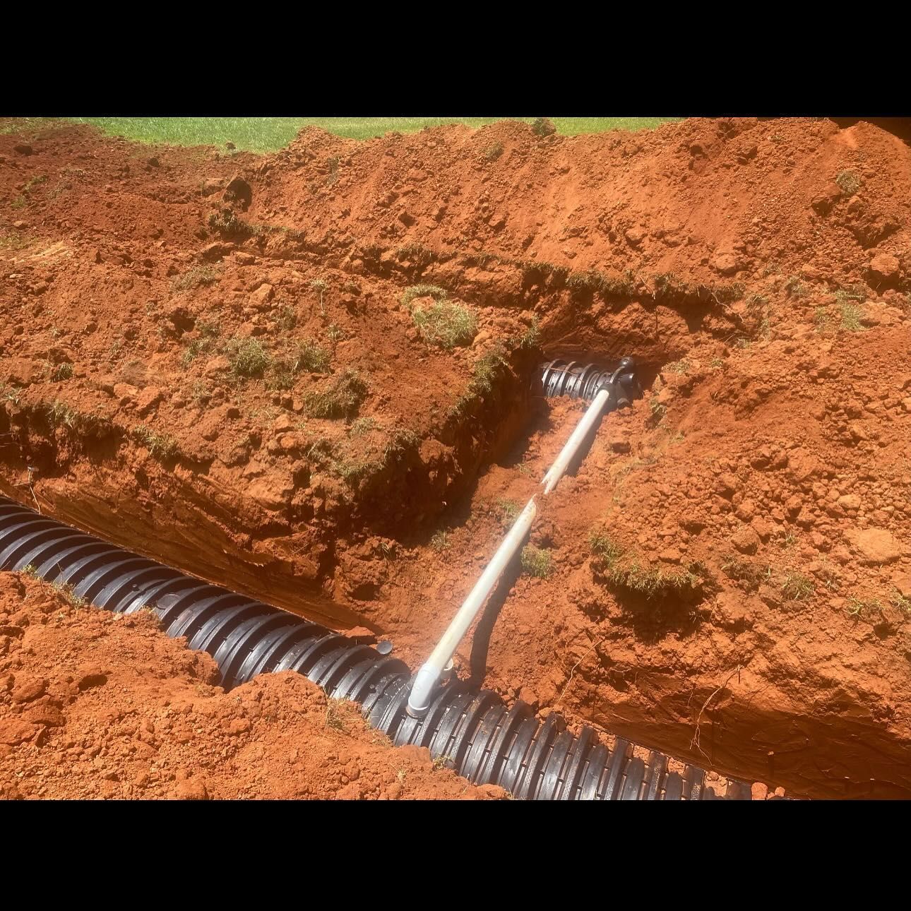 A trench with black corrugated pipe and a white handle tool, dug in red dirt, likely for drainage.