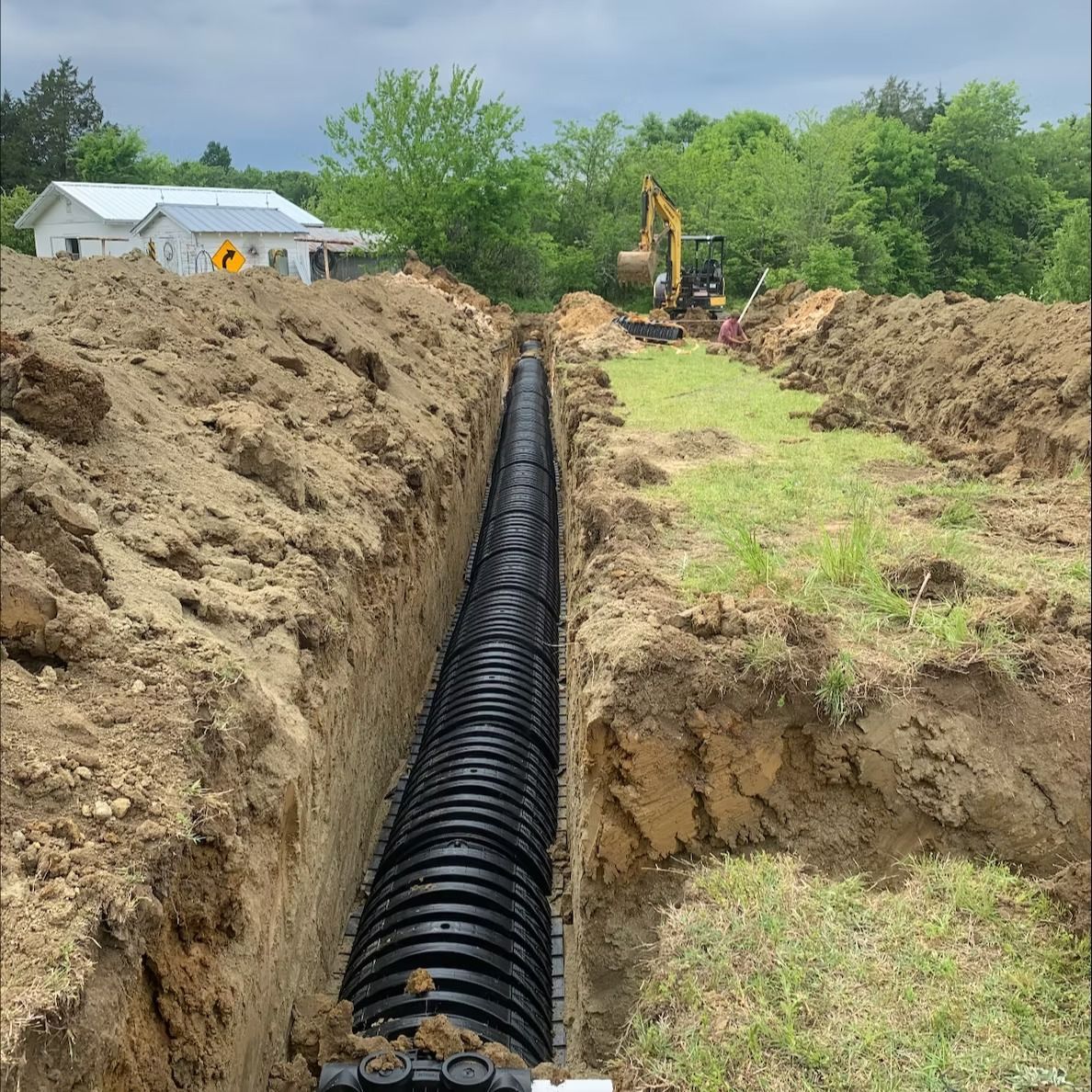 A long trench containing black drainage pipes, ready to be buried. A backhoe is visible in the background.