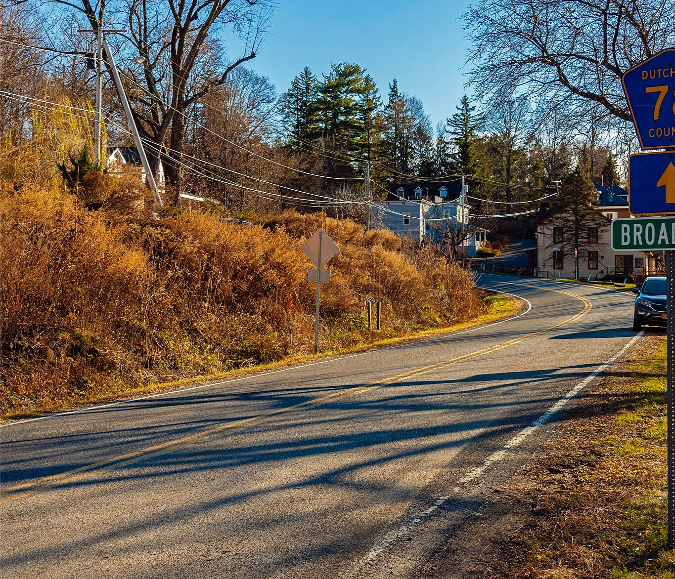 Road curving through a town, passing a sign for Broadway. Autumn foliage, blue sky.