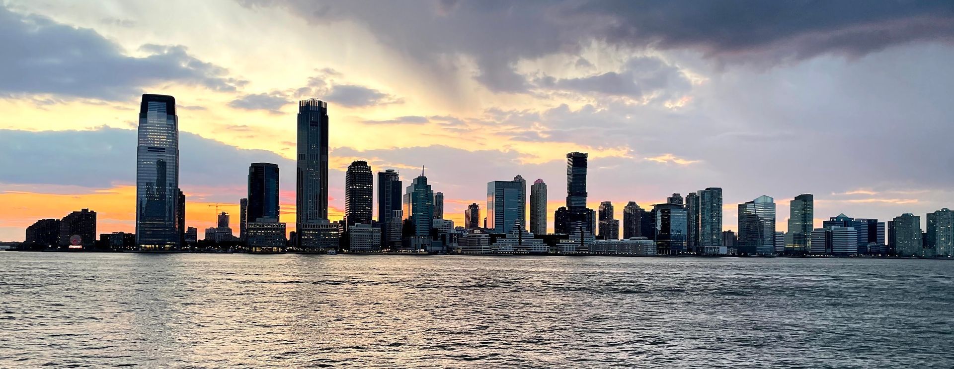 Skyline of city buildings reflecting in water at sunset.