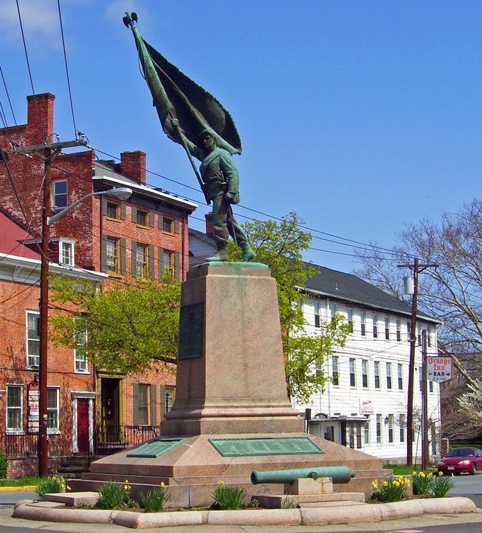 Civil War monument: soldier holding flag atop stone pedestal in city square. Buildings and blue sky in background.