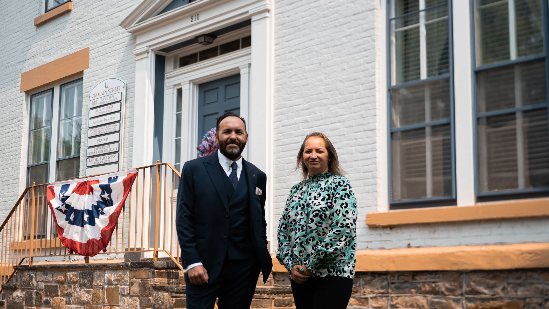 Two people stand in front of a white building with a banner. The man wears a suit and the woman a patterned top.