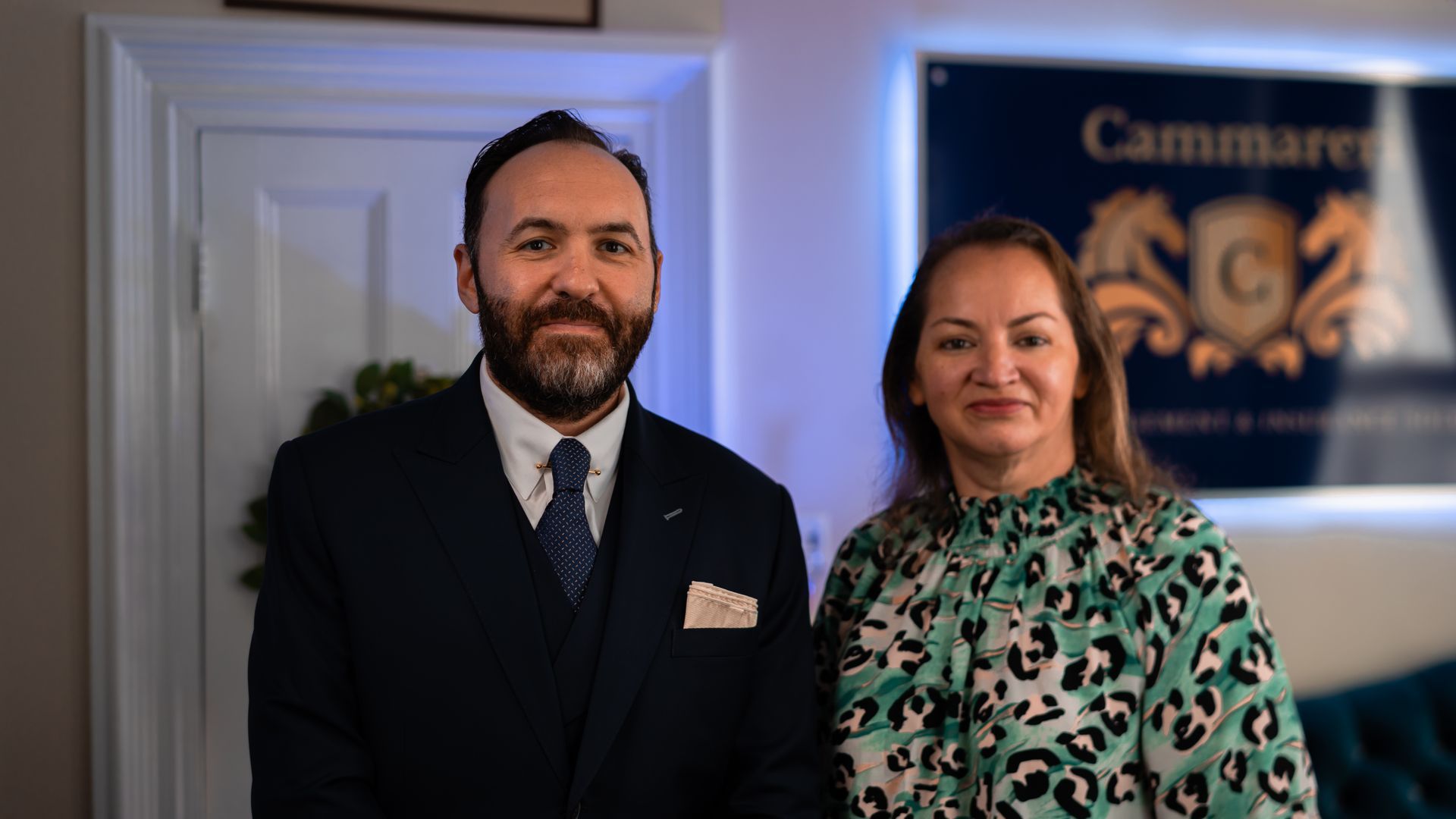 Man and woman in a professional setting, standing near a door and a sign with a logo.