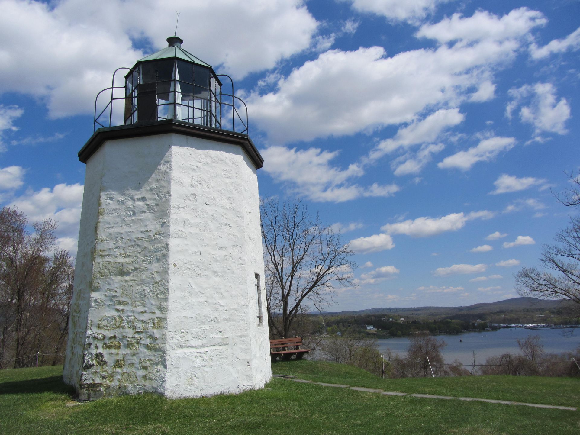 White, hexagonal lighthouse on a grassy hill; blue sky with clouds, water and distant shore in the background.