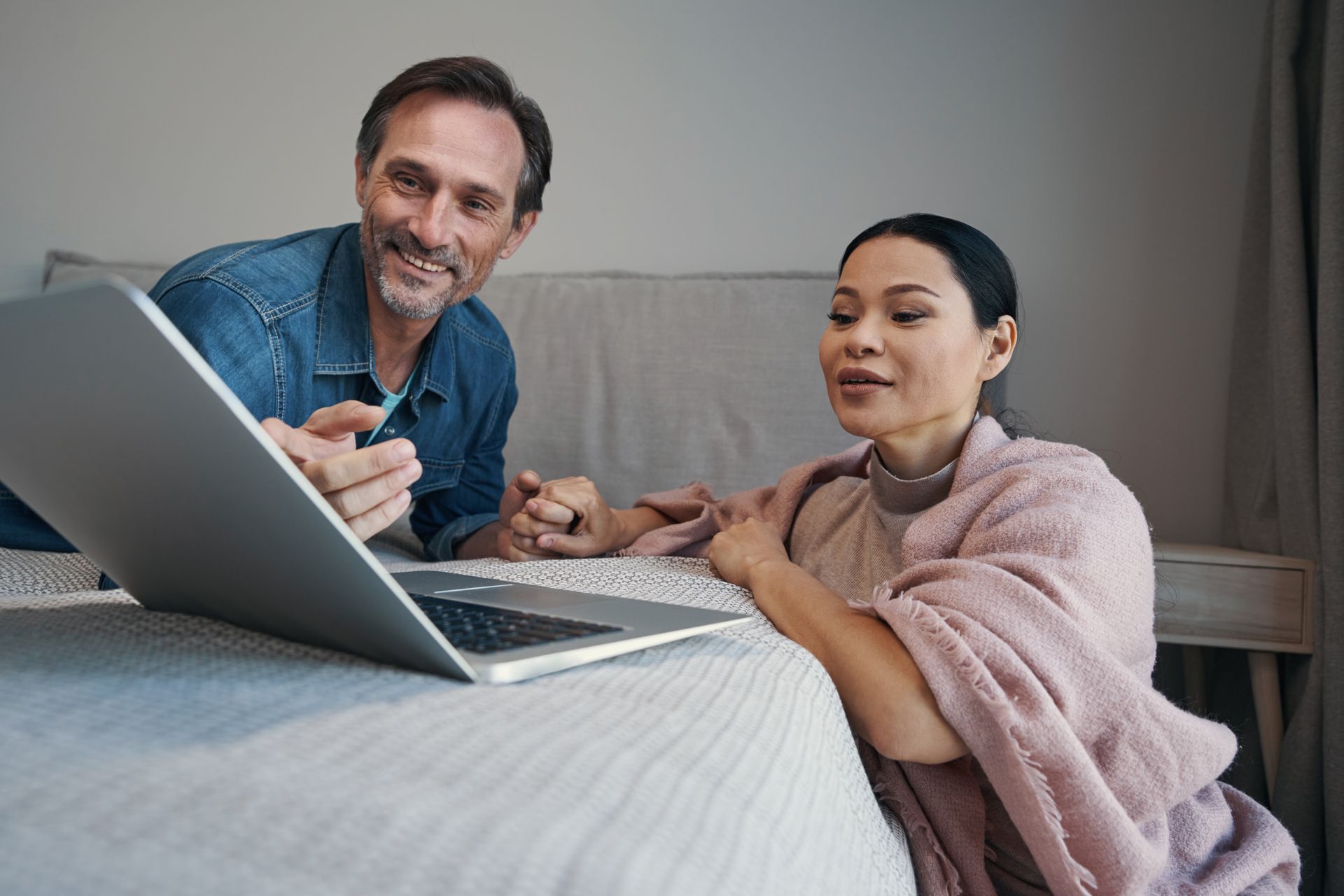 Couple looking at laptop screen, man pointing, woman wrapped in a blanket, indoor setting.