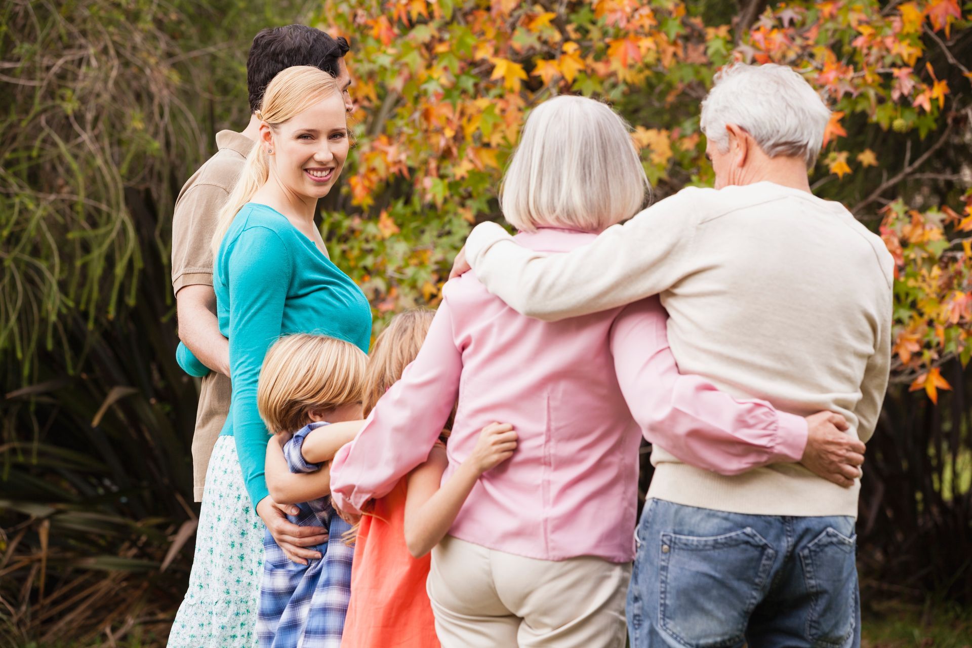 Family with arms around each other in a park. Blonde woman smiles at camera, fall foliage in background.