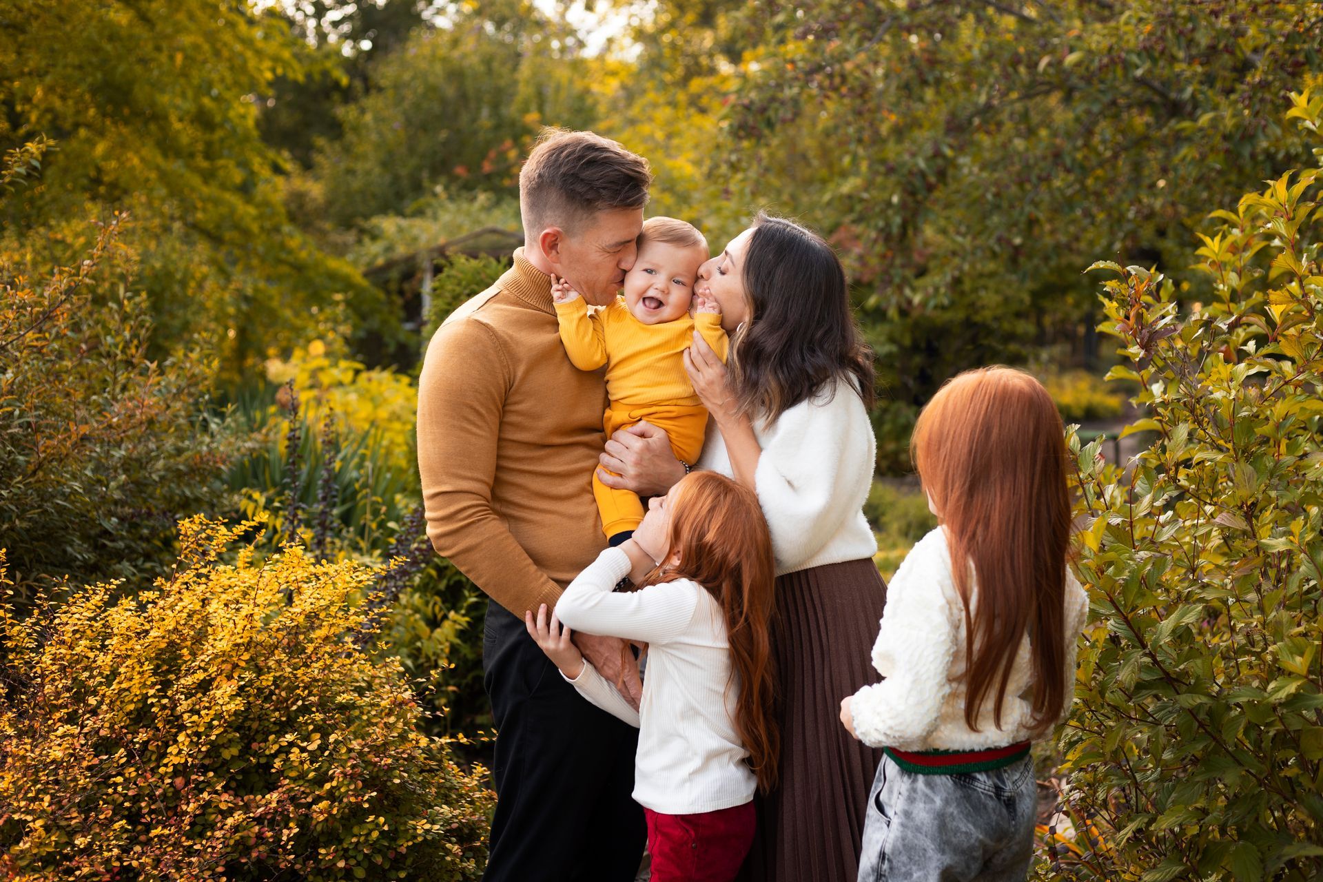 Family of five in a park, parents kissing a child, two girls nearby, surrounded by greenery.