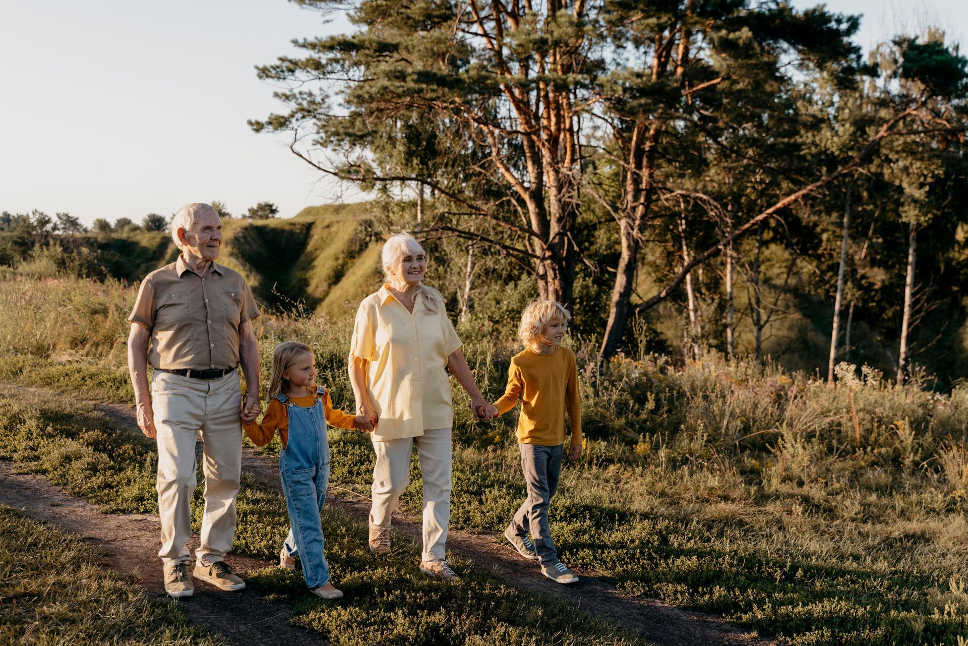 Two older adults and two children hold hands while walking on a path outdoors.