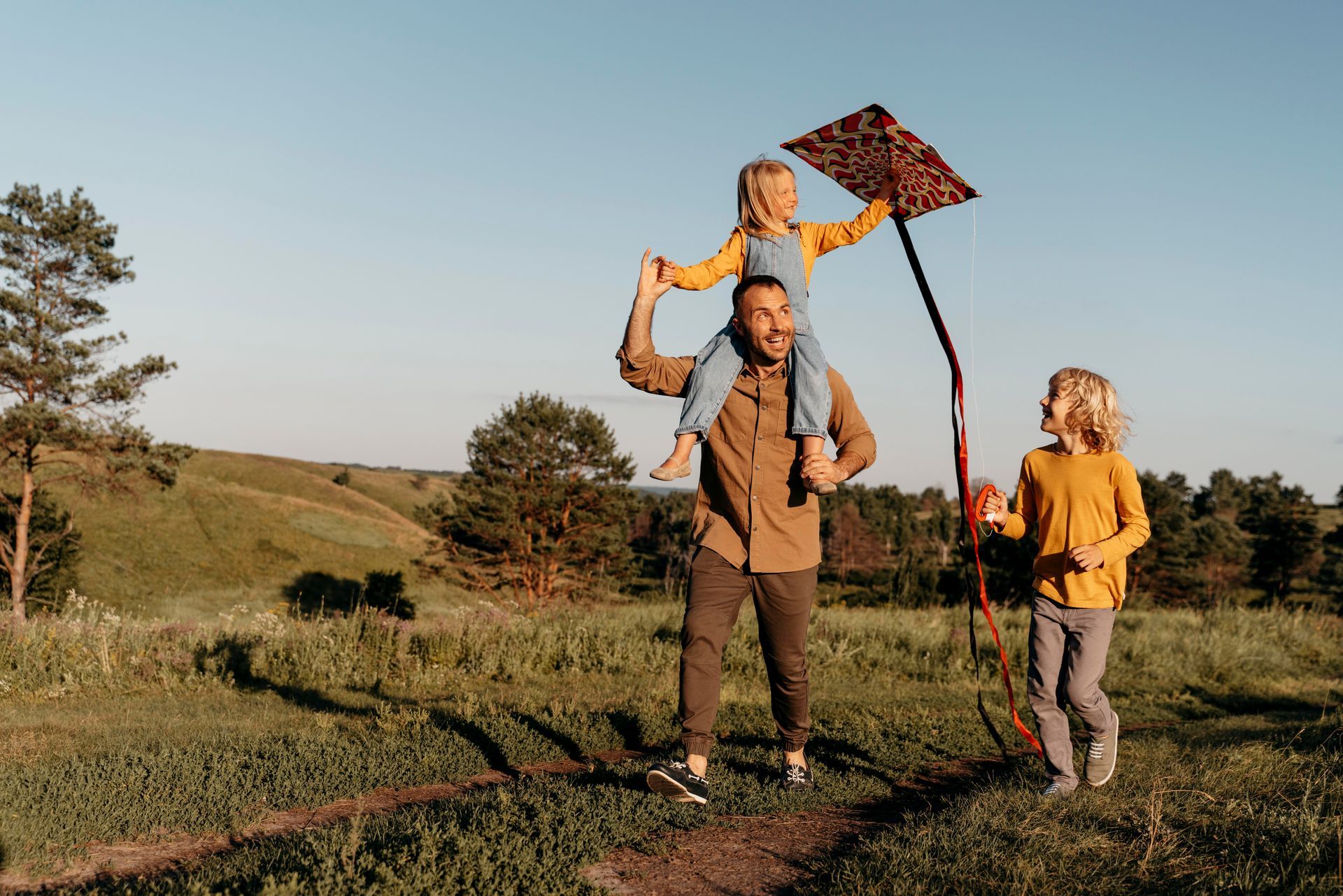 Father carrying child on his shoulders, flying kite with another child in a sunny field.