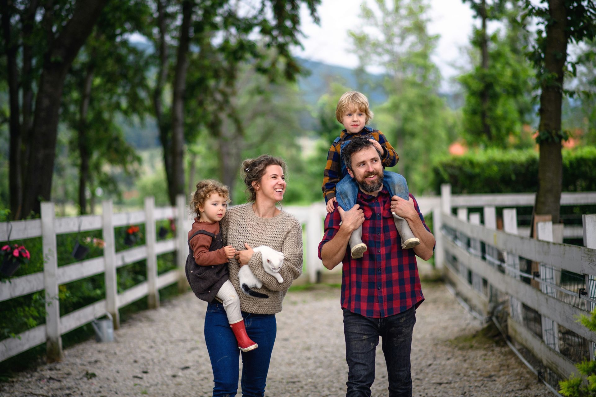 Family walking on a path: mother carrying child with lamb, father carrying child on shoulders, white fence and trees.