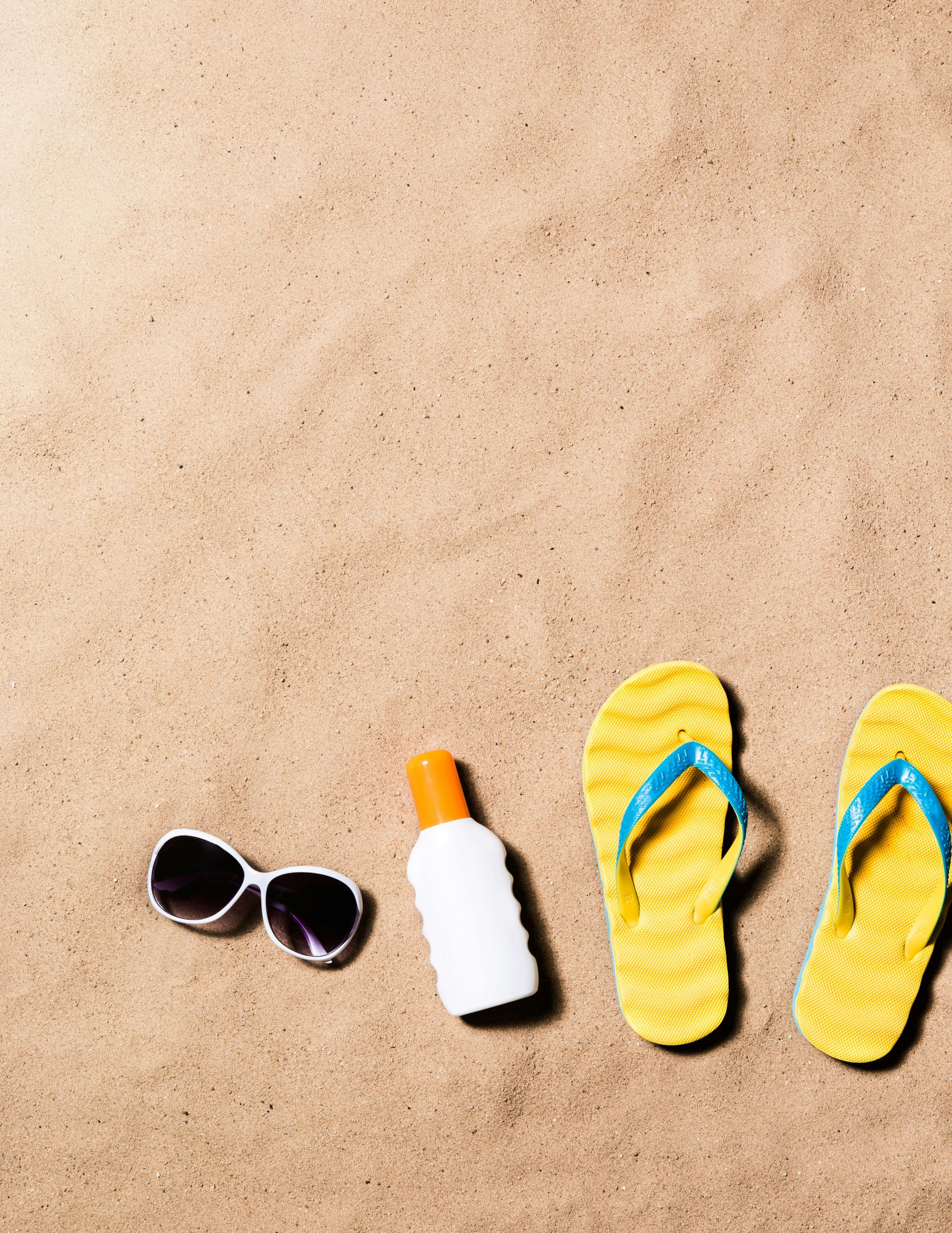 Sunglasses, sunscreen, and yellow flip-flops on sandy beach.