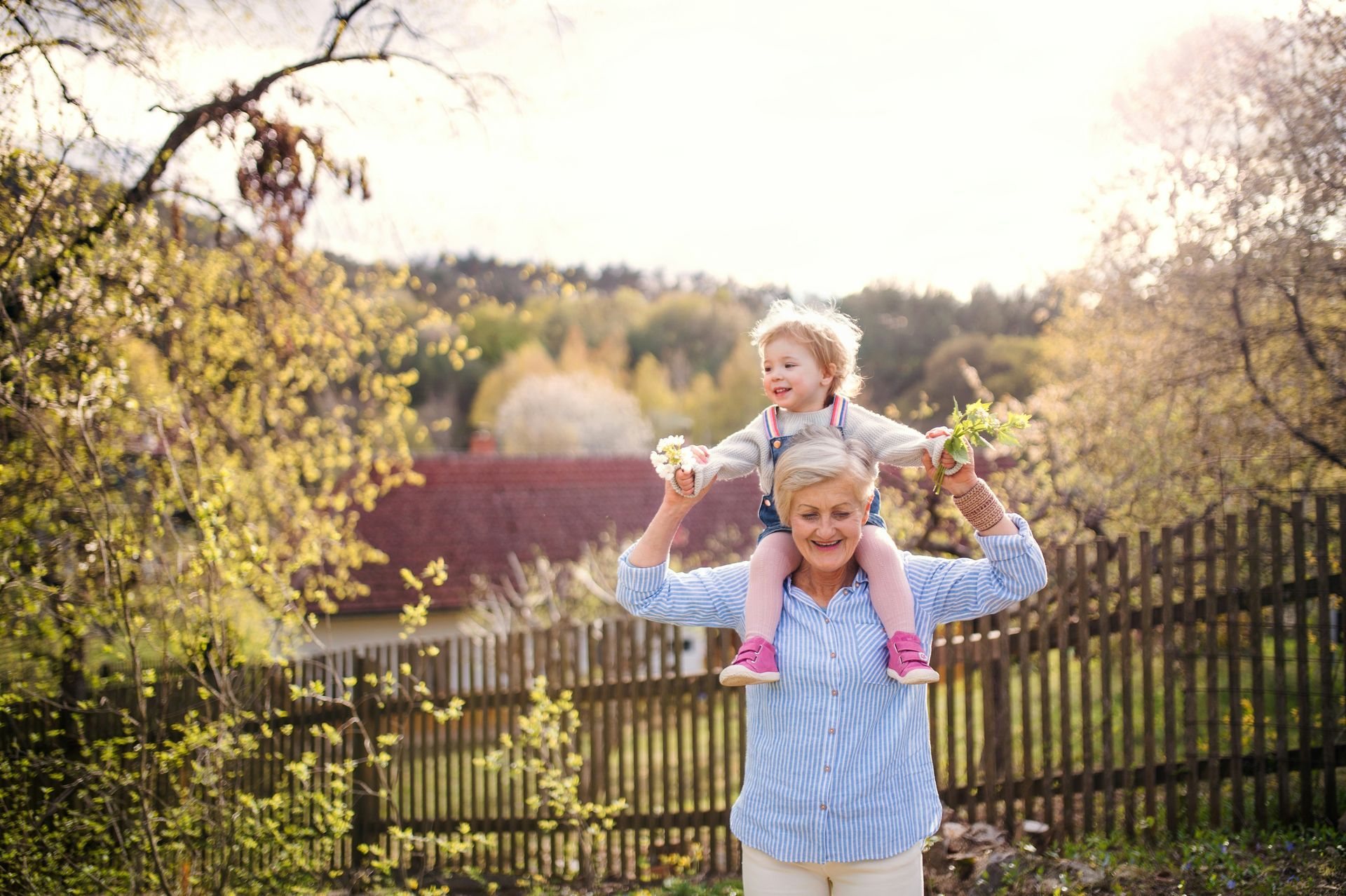Woman carrying a child on her shoulders outdoors near a wooden fence, smiling.