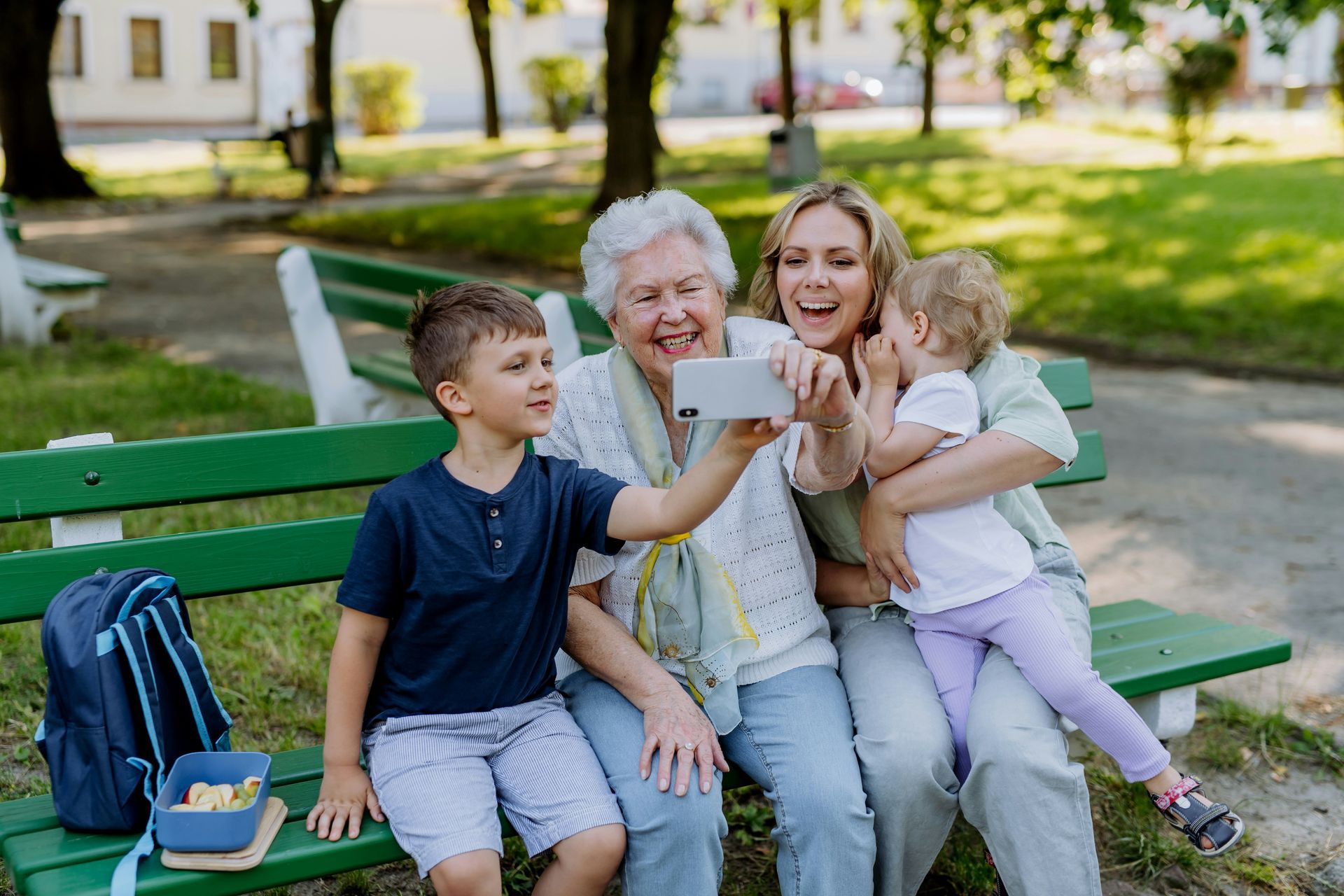 Family of four smiling, taking a selfie on a park bench. A backpack and a snack container sit nearby.