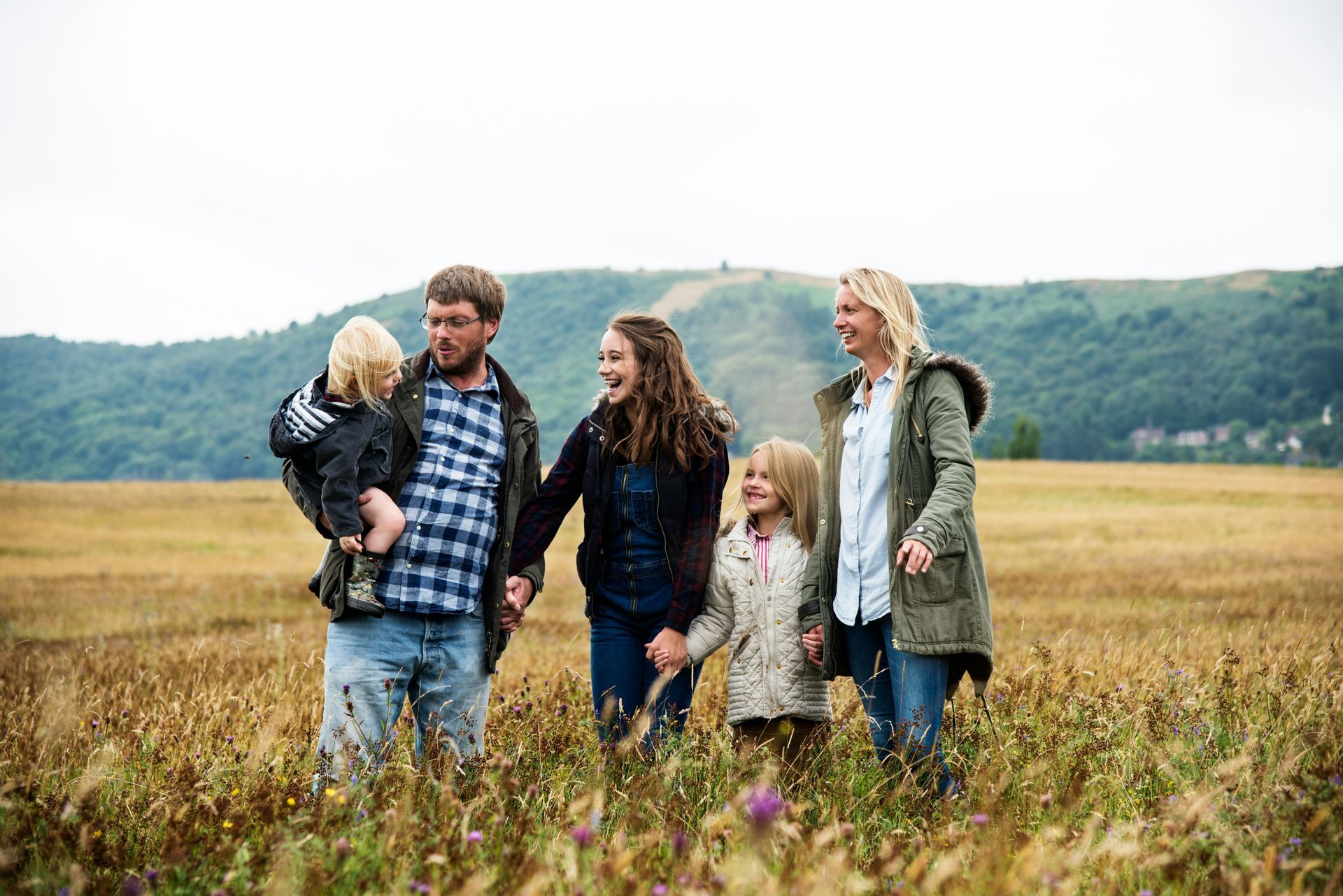 Family of five walking through a field, holding hands, smiling. Mountains in the background.