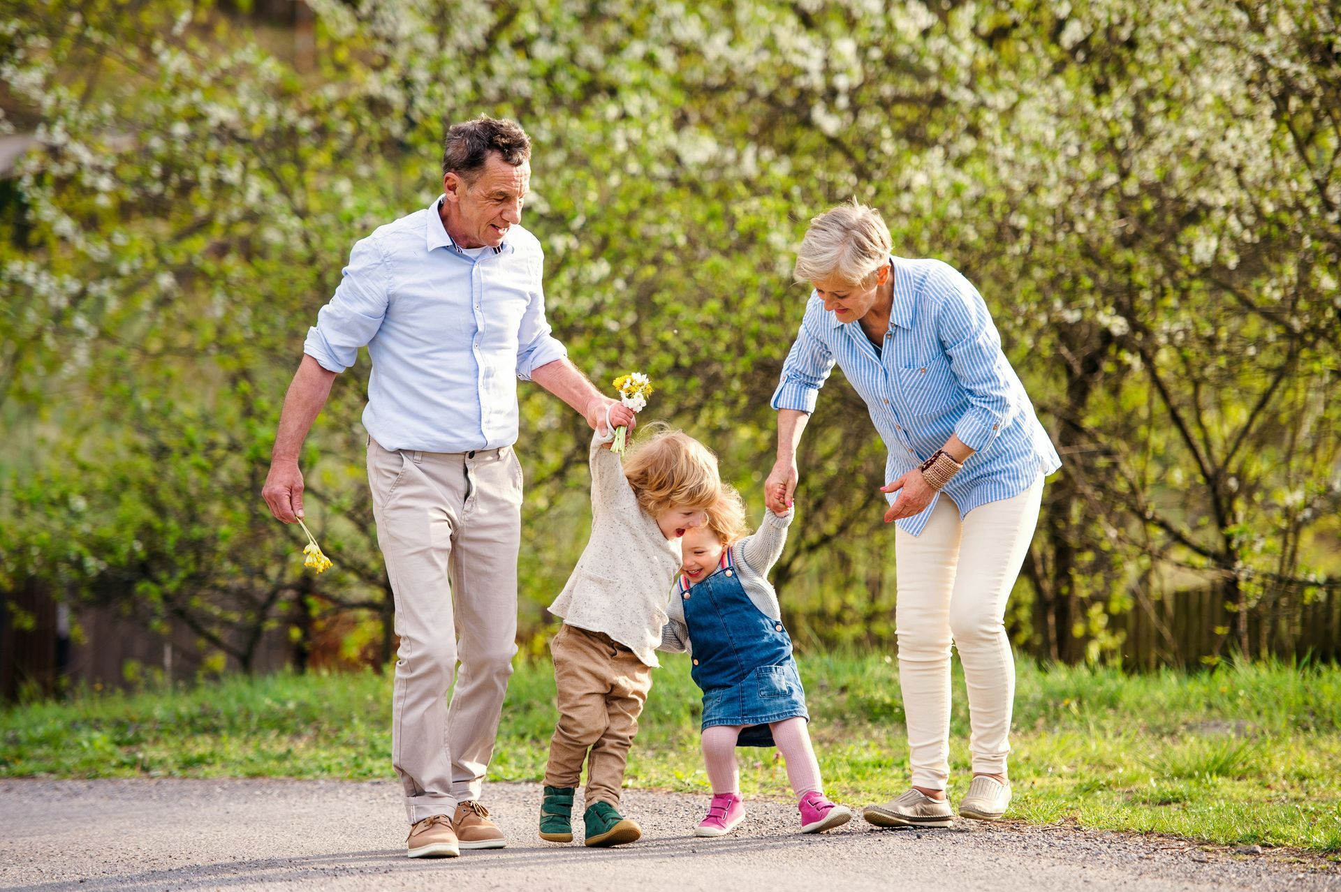 Grandparents helping two young children walk on a path in a park; sunny, happy smiles.