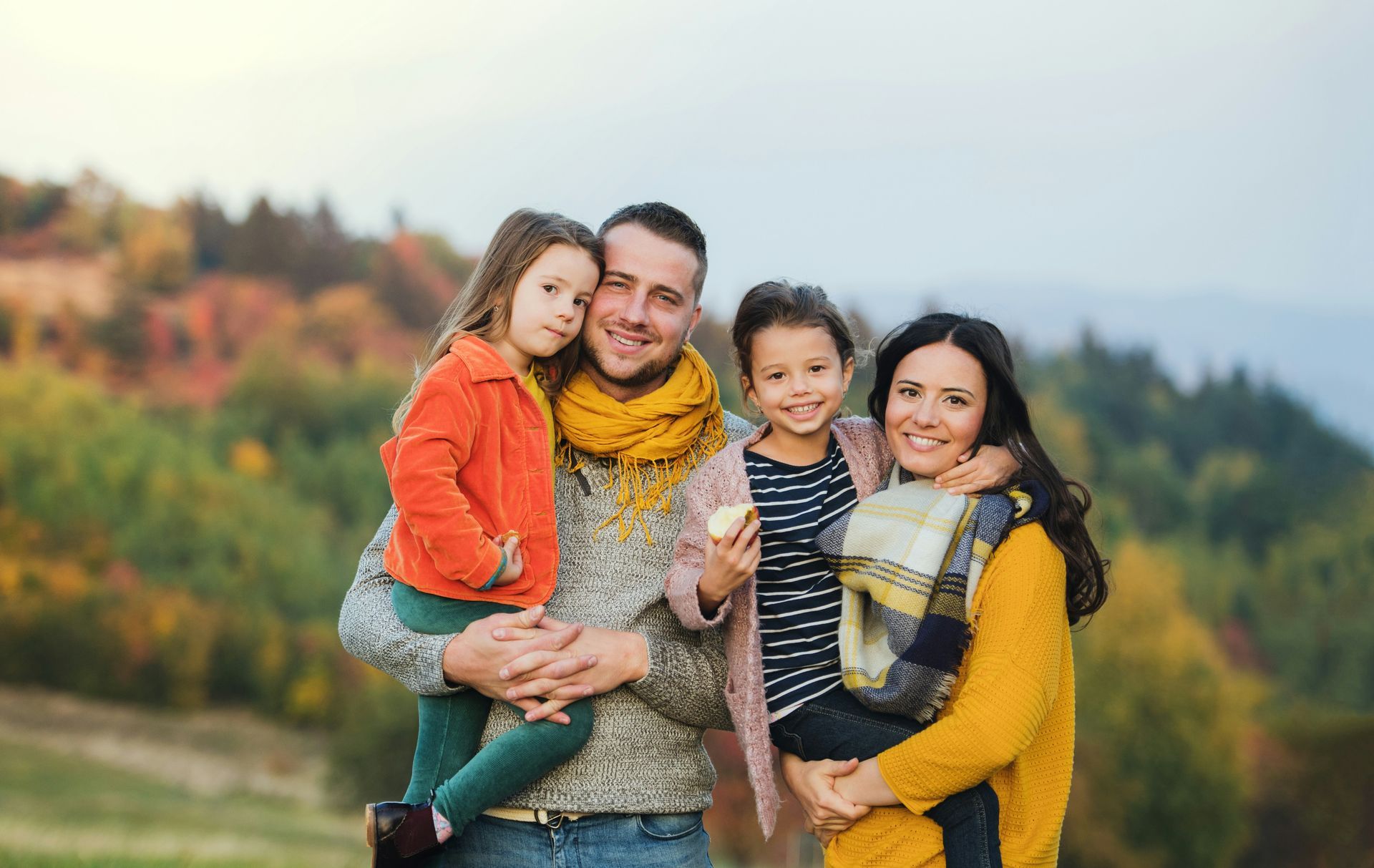 Family of four posing outdoors with fall foliage; children being held.