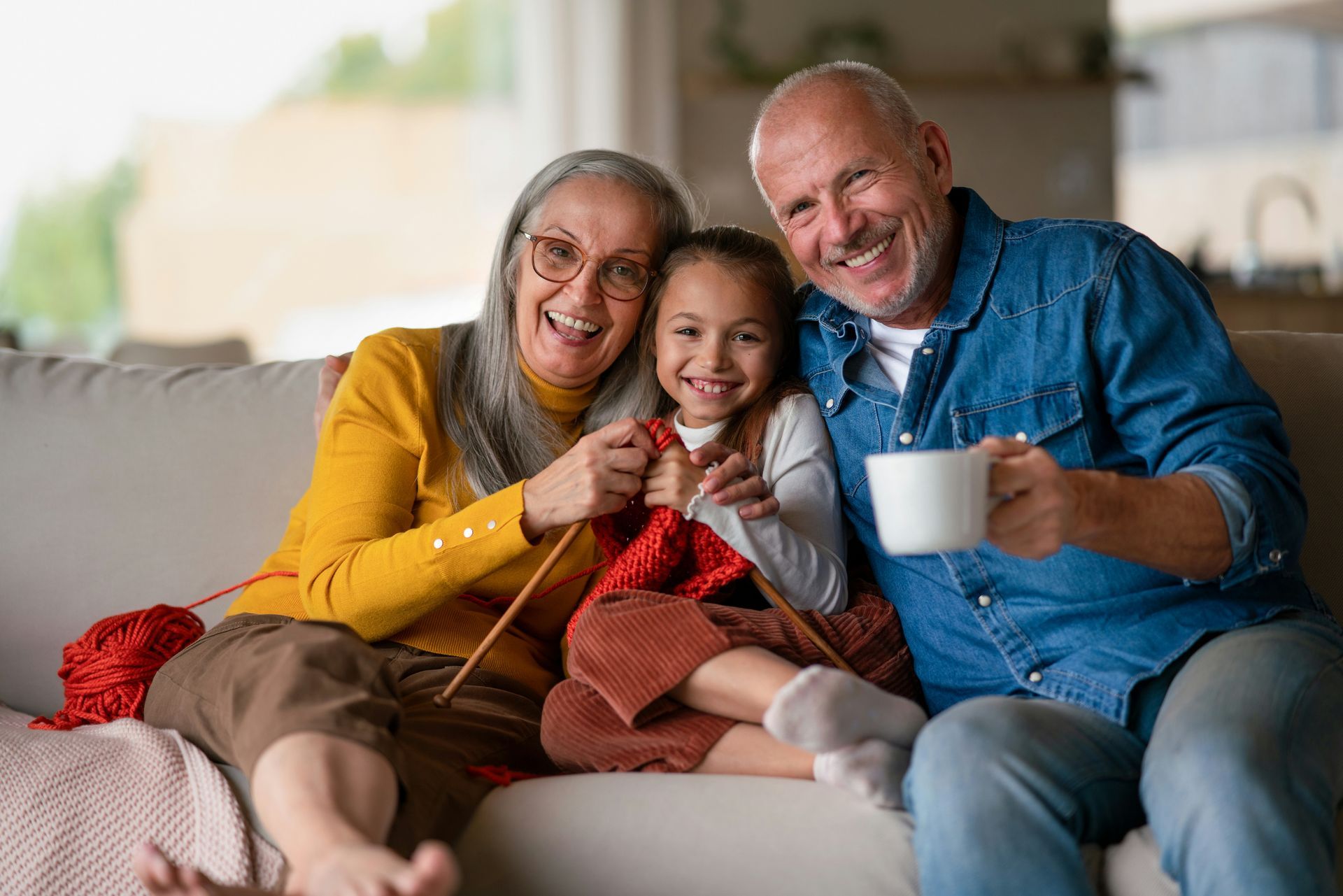 Grandparents and grandchild smiling on a couch; woman knitting with red yarn, man holding a mug.