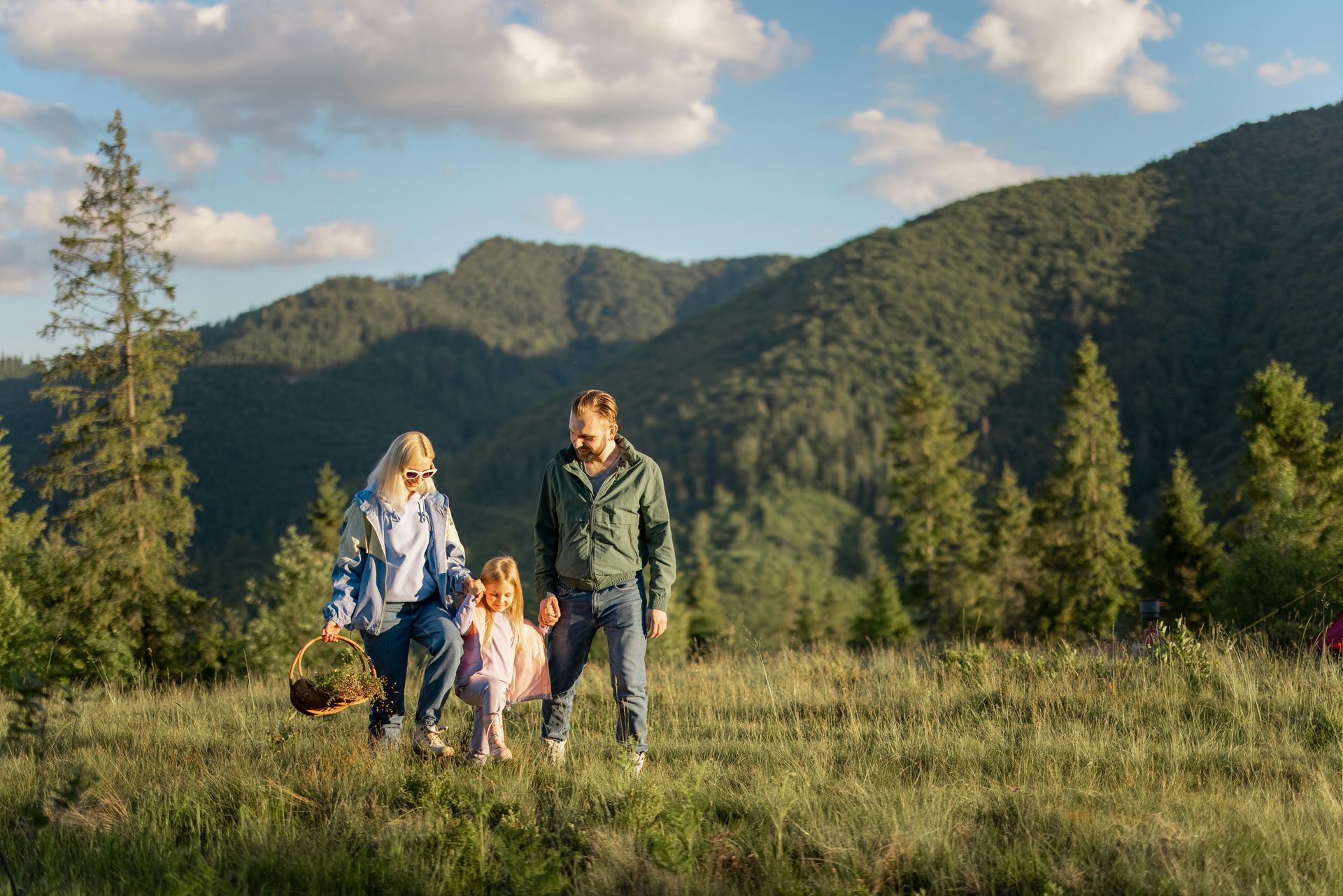 Family walking in a mountain meadow; woman holding basket, man and child holding hands; trees, sky.