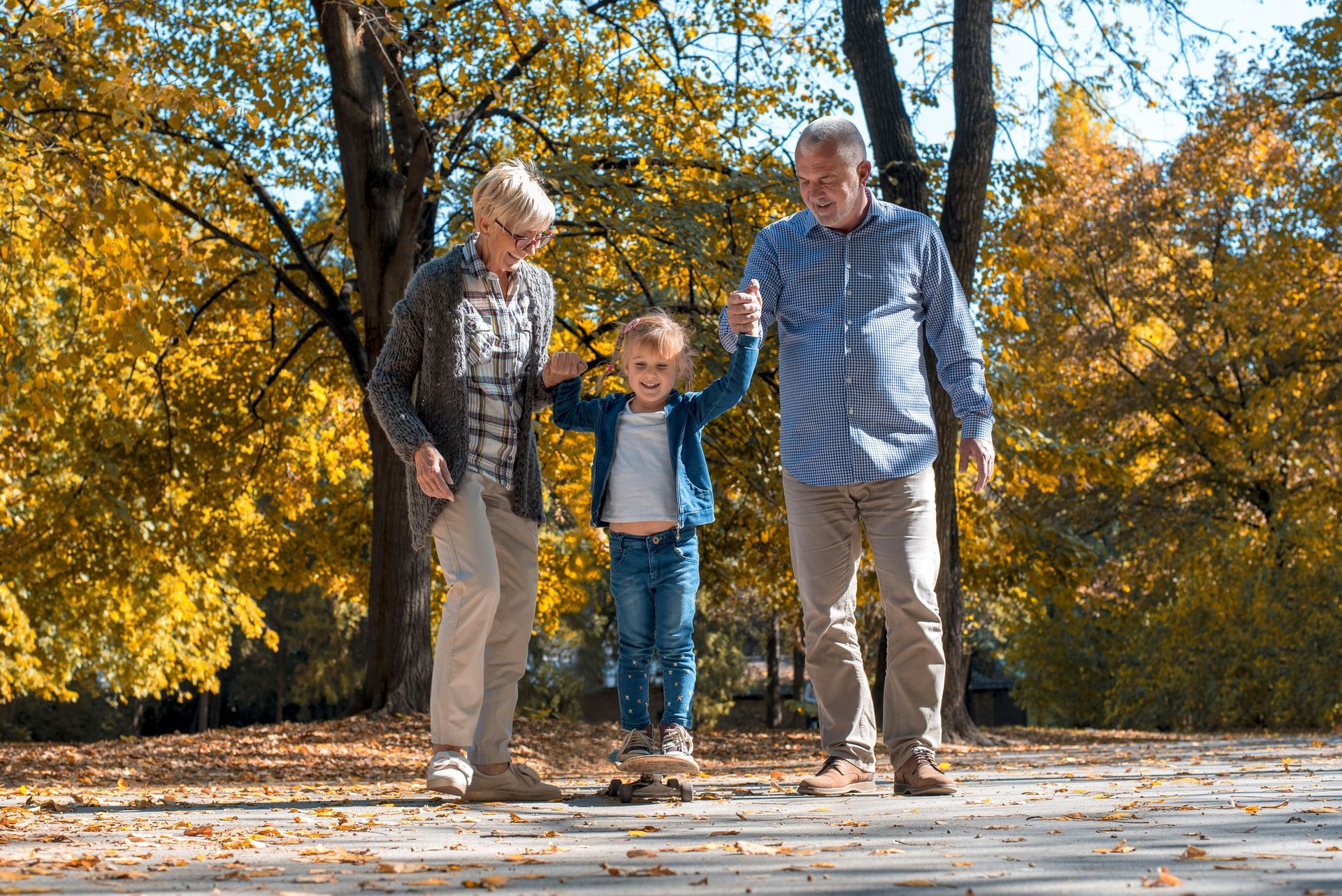 Grandparents hold hands with a child, walking outdoors, autumn leaves in the background.