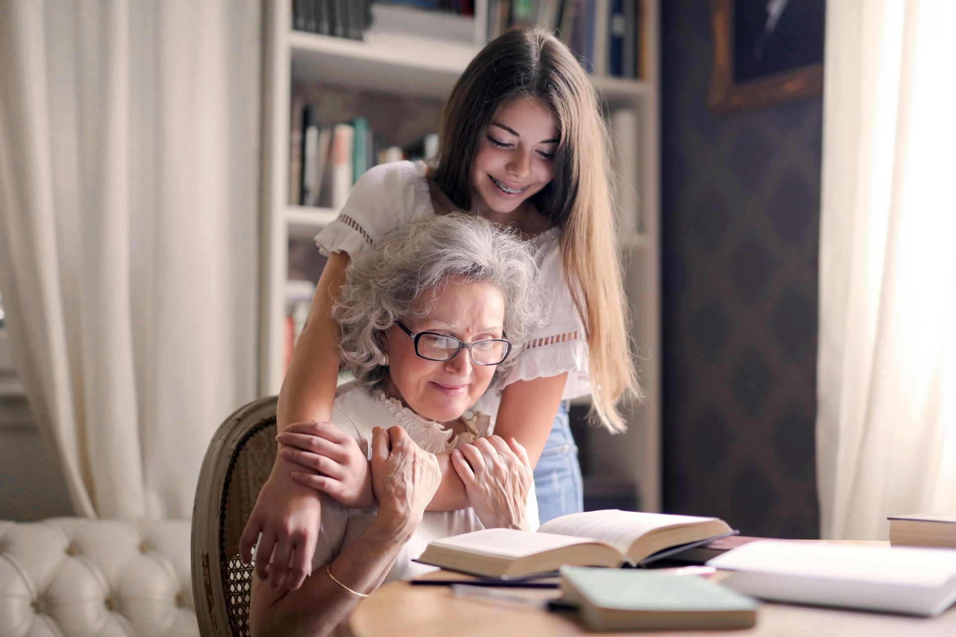 Woman embraces older person seated at table with books.