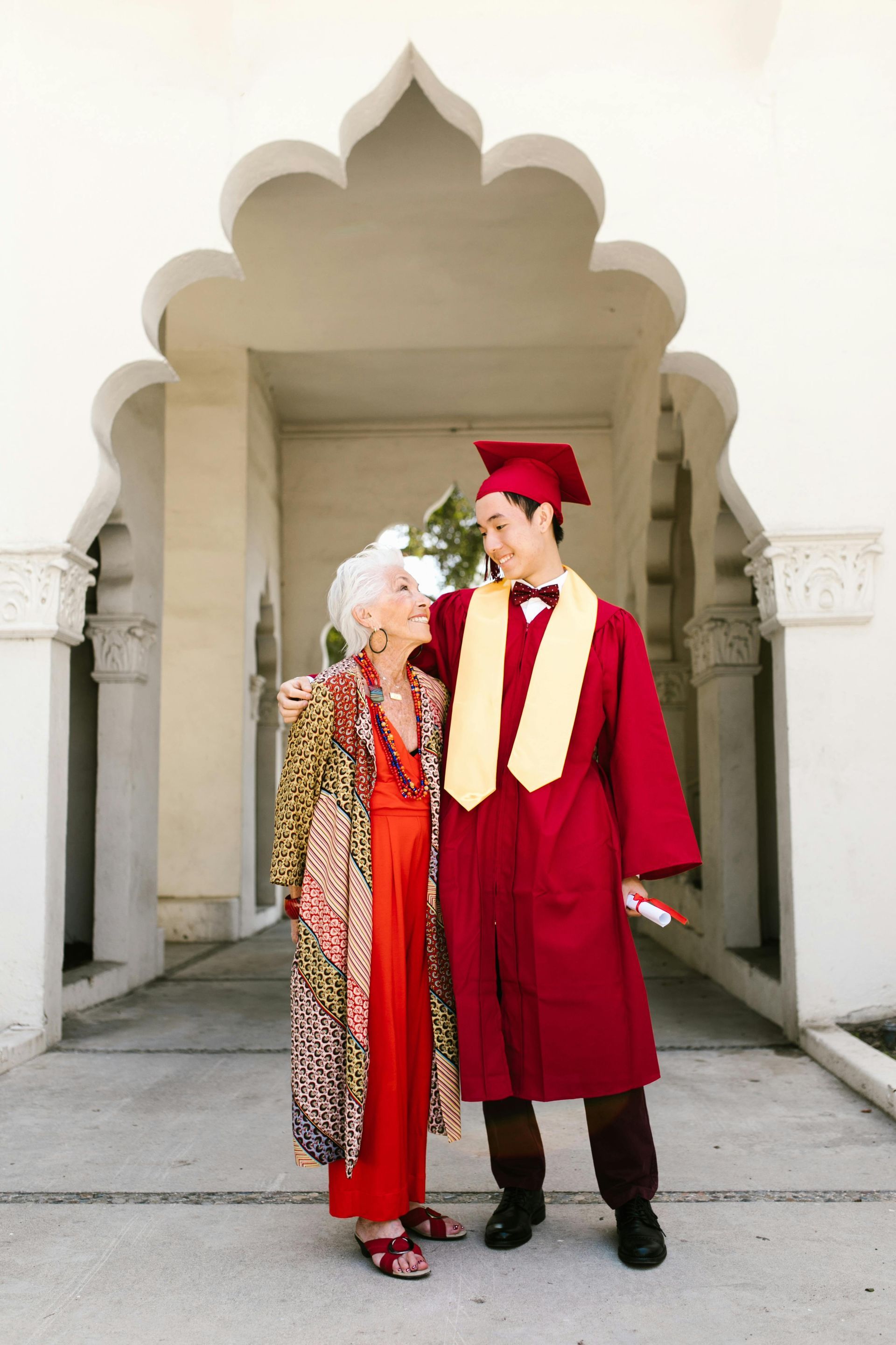 A graduate in red gown and cap poses with an older person in a patterned coat. They stand under an archway.