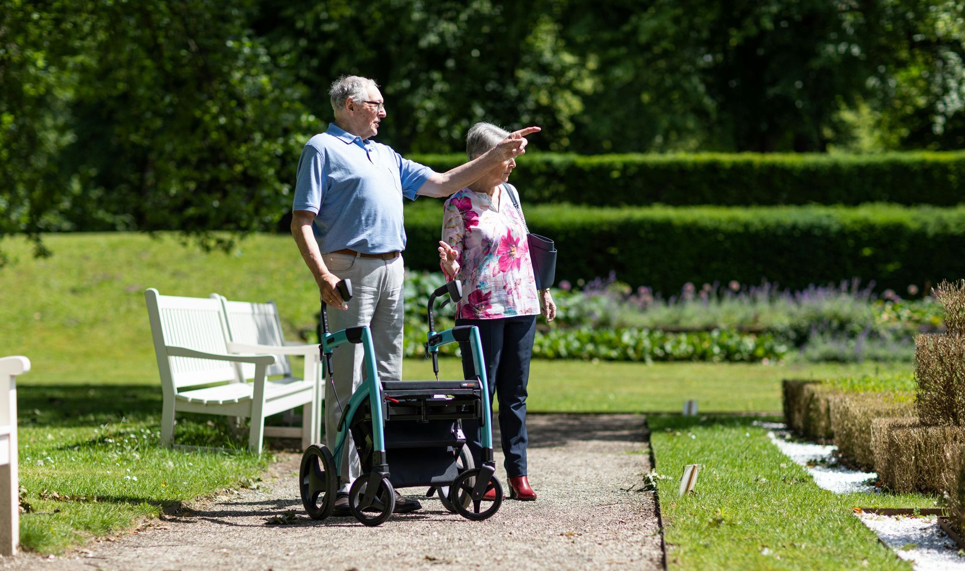 Elderly couple in a garden, one pointing, with a rolling walker on a path next to trimmed bushes.