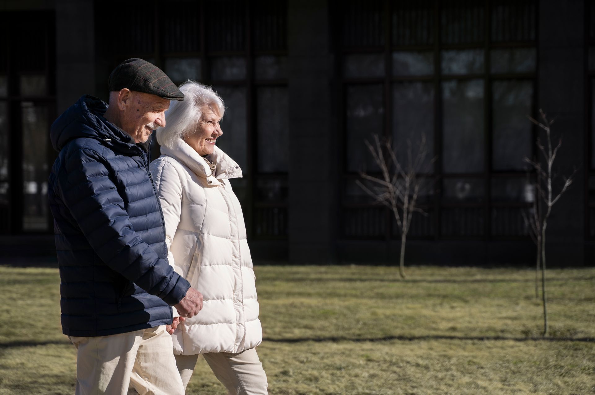 Couple strolling outdoors, holding hands. Man in blue jacket, woman in white coat. Sunny day.