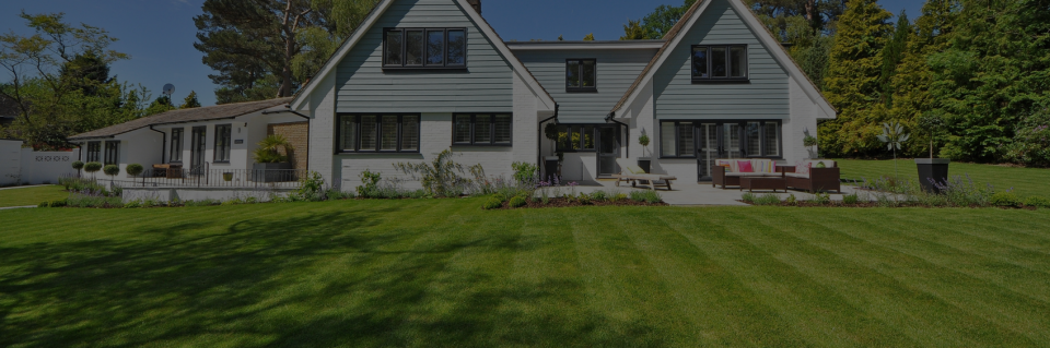 A two-story house with white siding and gray shingles sits behind a wide, mowed lawn on a sunny day.