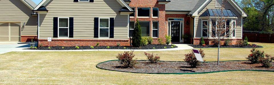 A suburban house with beige siding, a brick lower facade, and a landscaped yard featuring a central shrubbed island.
