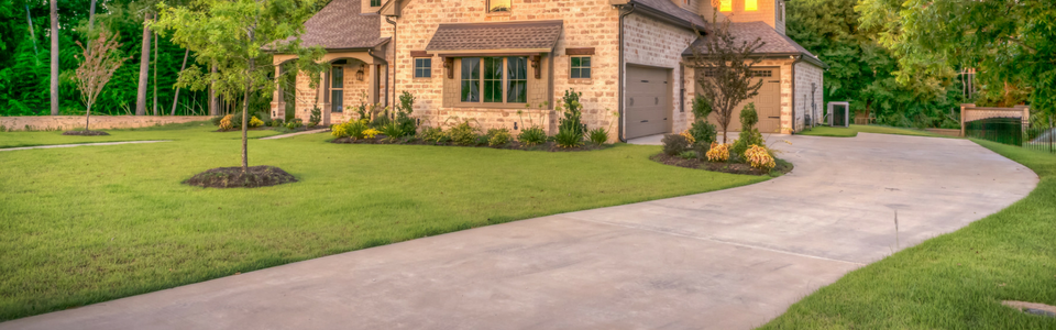 A stone house with a multi-car garage, manicured lawn, and a wide concrete driveway leading to the street.