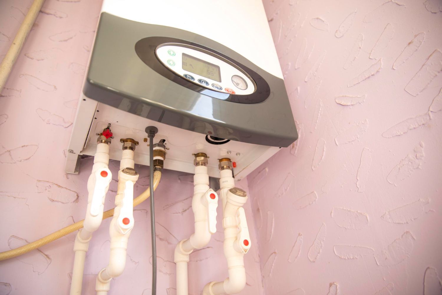 A boiler with pipes attached to it is hanging on a pink wall.