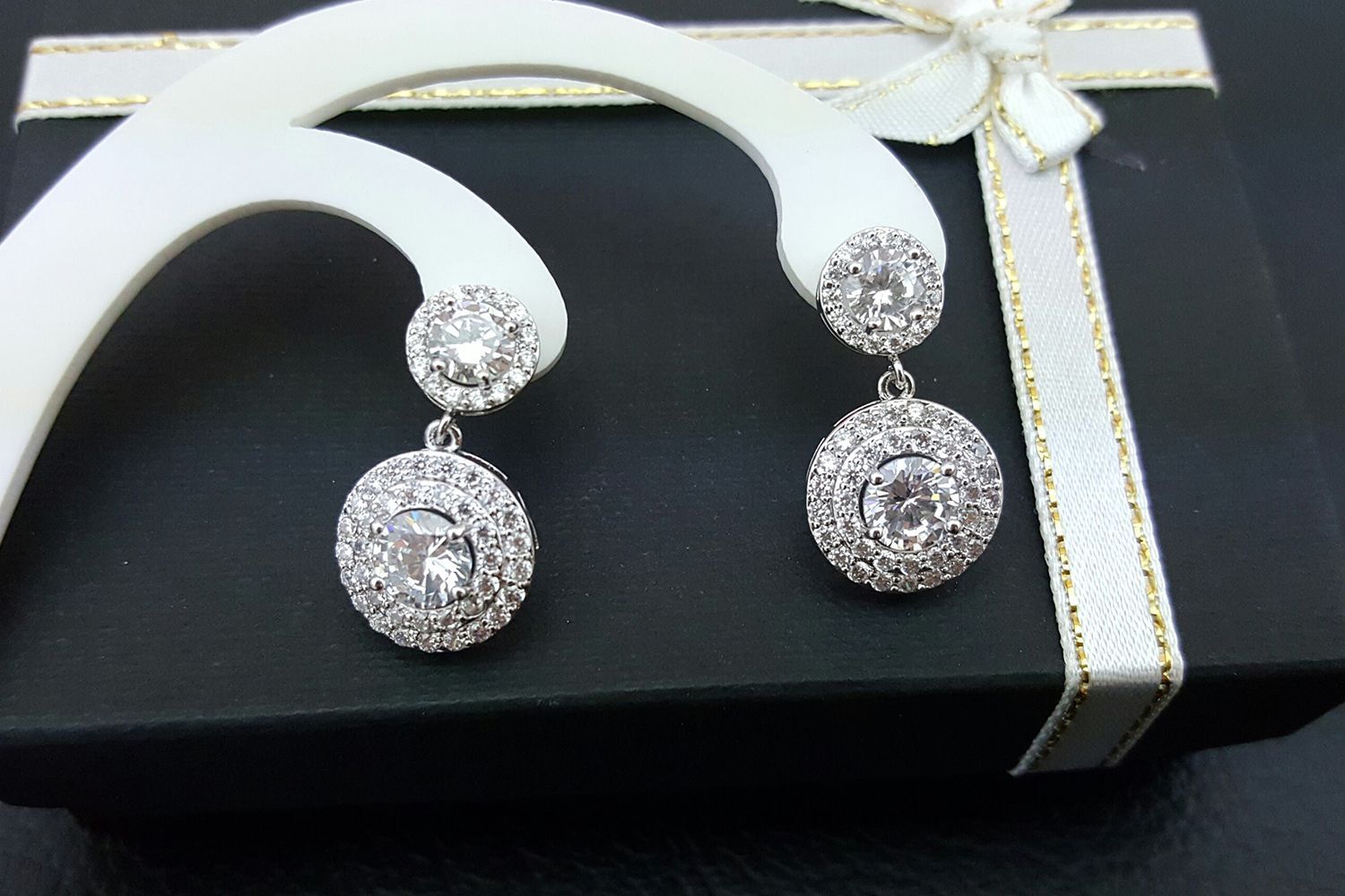Silver earrings with clear stones, dangling on a white stand, atop a black box with ribbon.