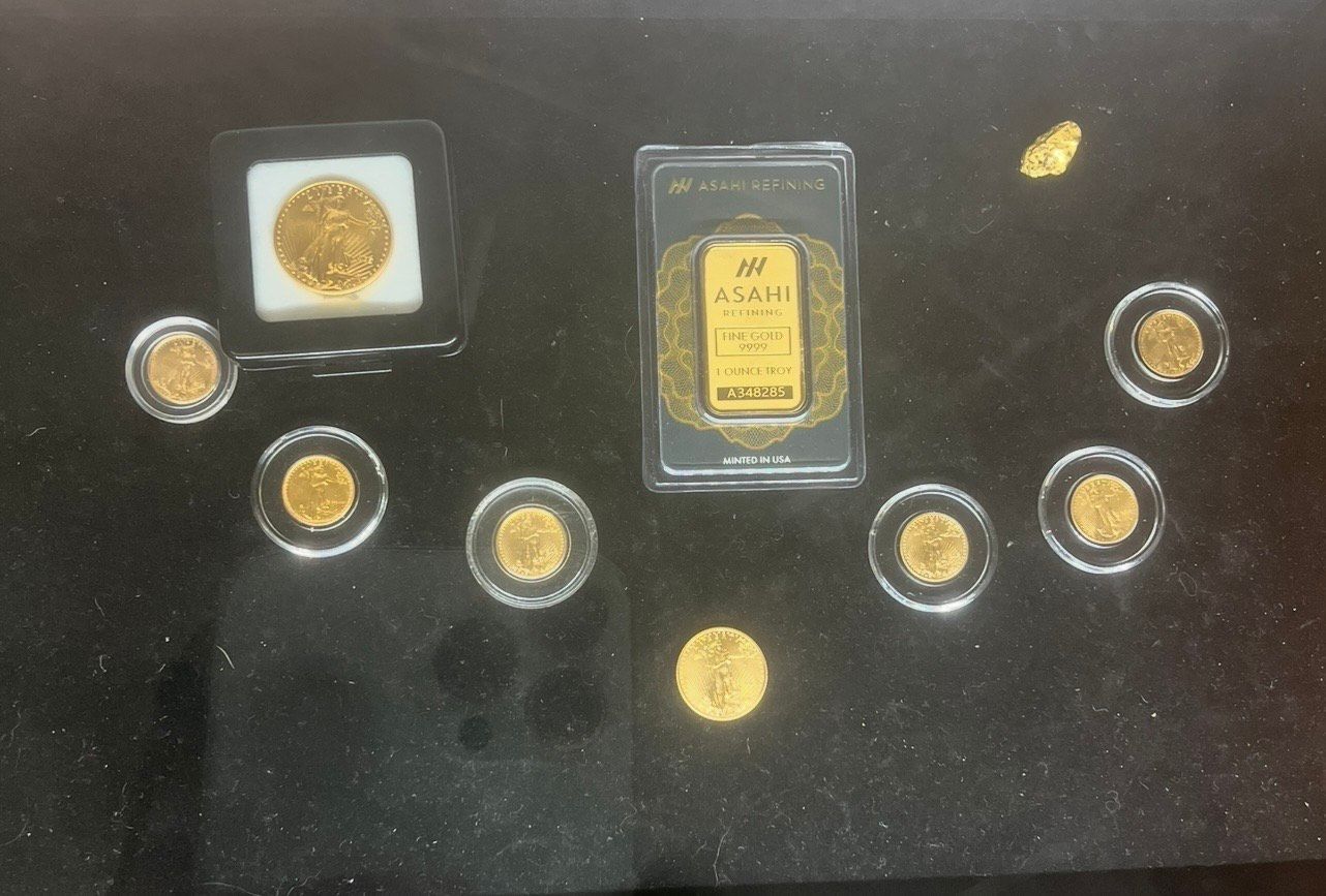 Gold jewelry display in a shop, featuring necklaces and bracelets on mannequins and in cases. Gold coins and a gold bar displayed on a dark surface.