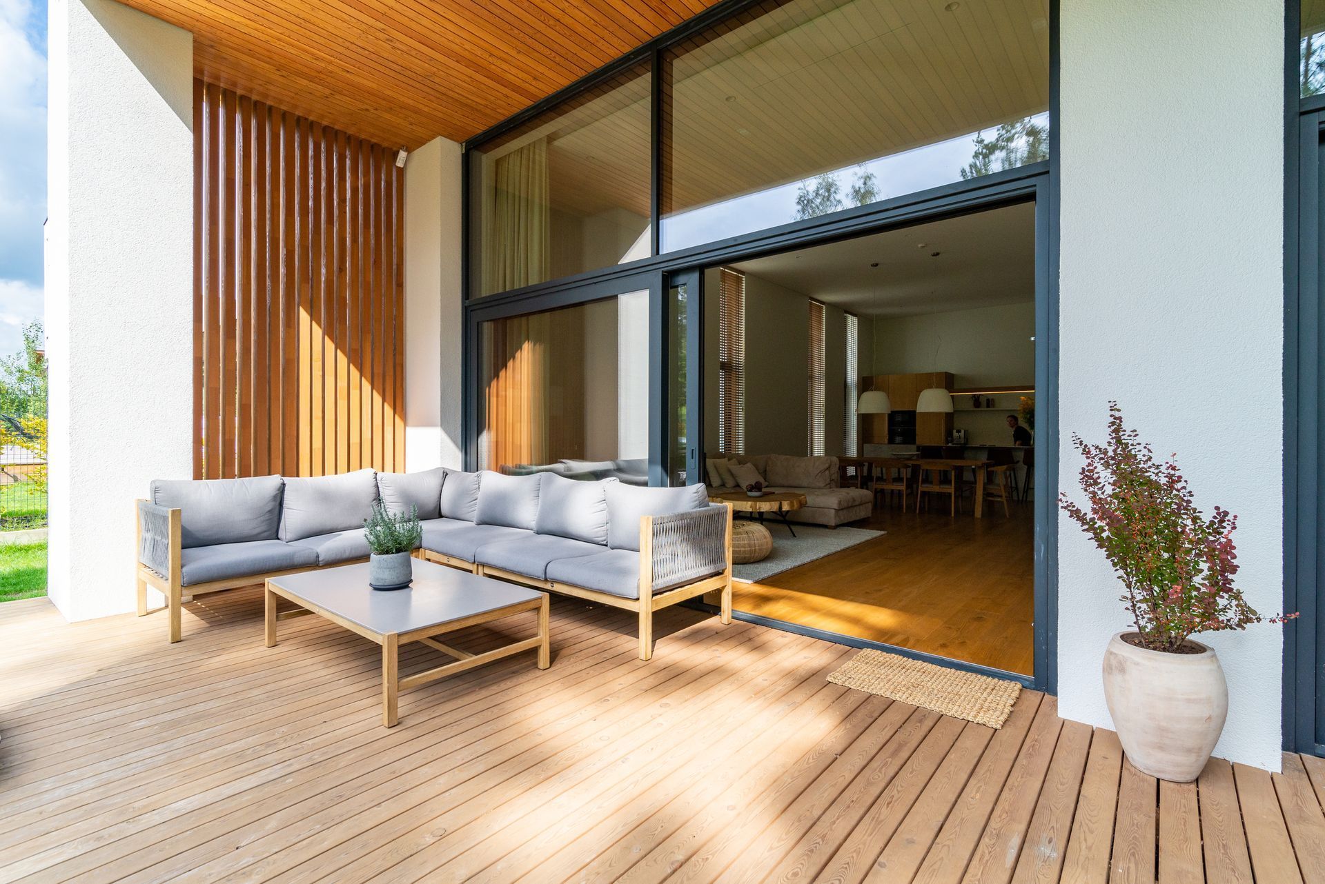 Modern patio with gray outdoor sofa, wooden deck, and open sliding doors to a bright living room