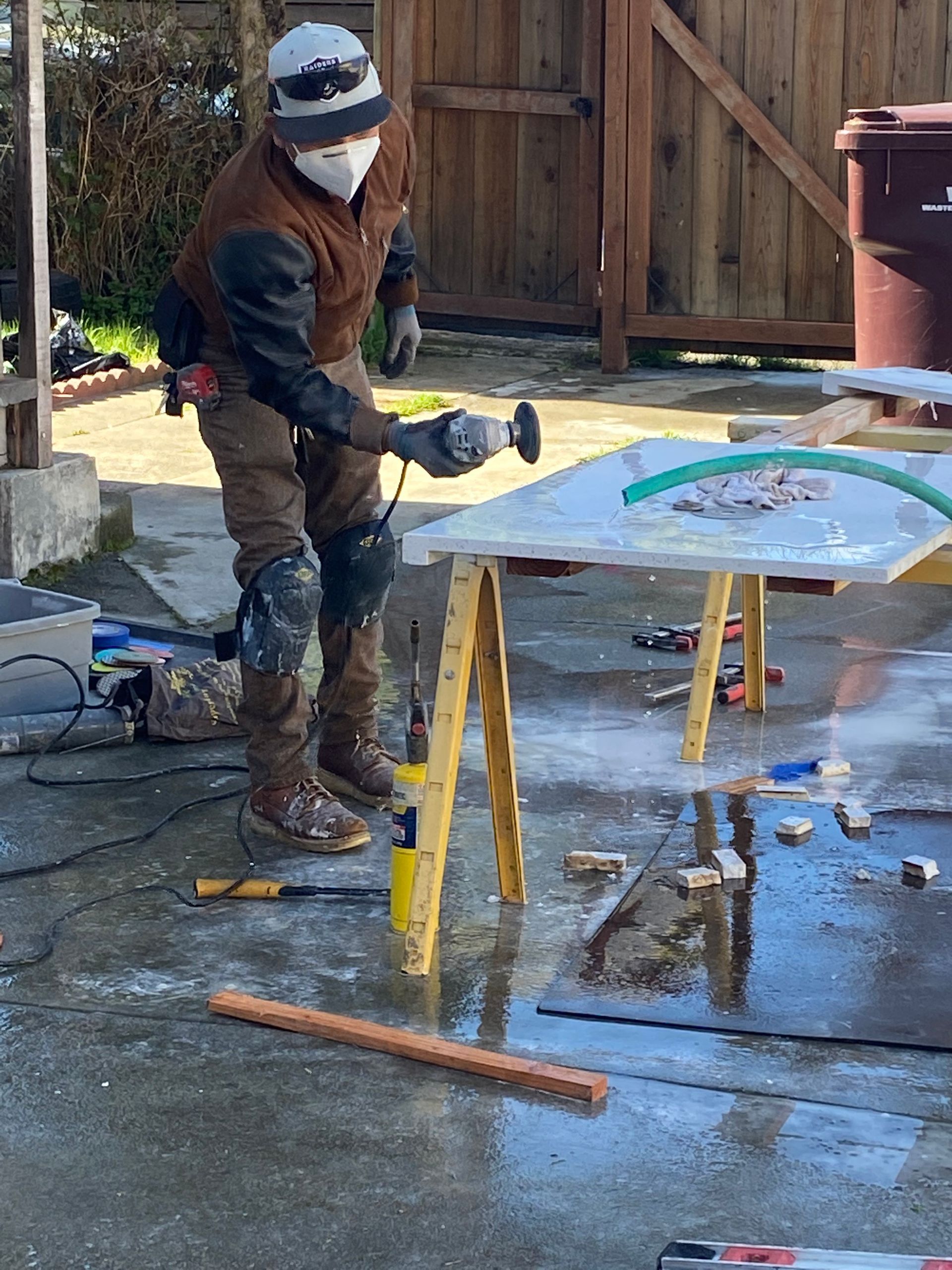 Person in winter gear washing a colorful tabletop outdoors with a hose, standing on a wet patio.