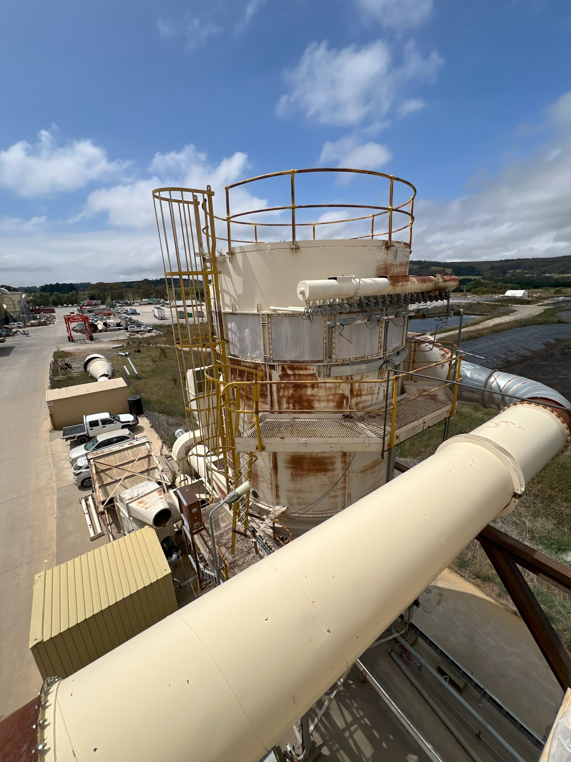Tall, beige industrial structure with yellow railings and ladder, against a cloudy sky and industrial setting.