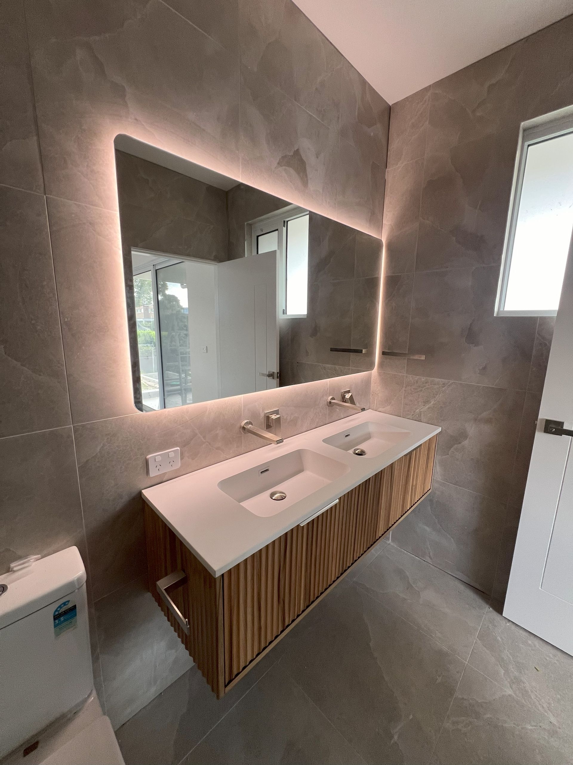 Modern bathroom with a floating wood vanity, double sinks, and a backlit mirror. Grey tile walls and floor.
