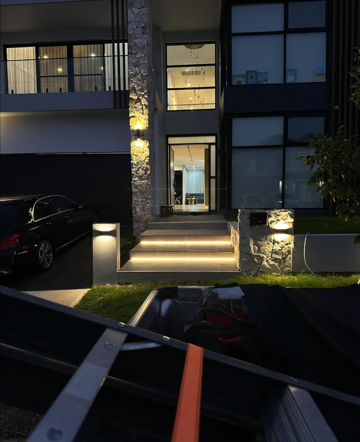 Night exterior of a modern home with lit pathway and front door. A car is parked on the left.