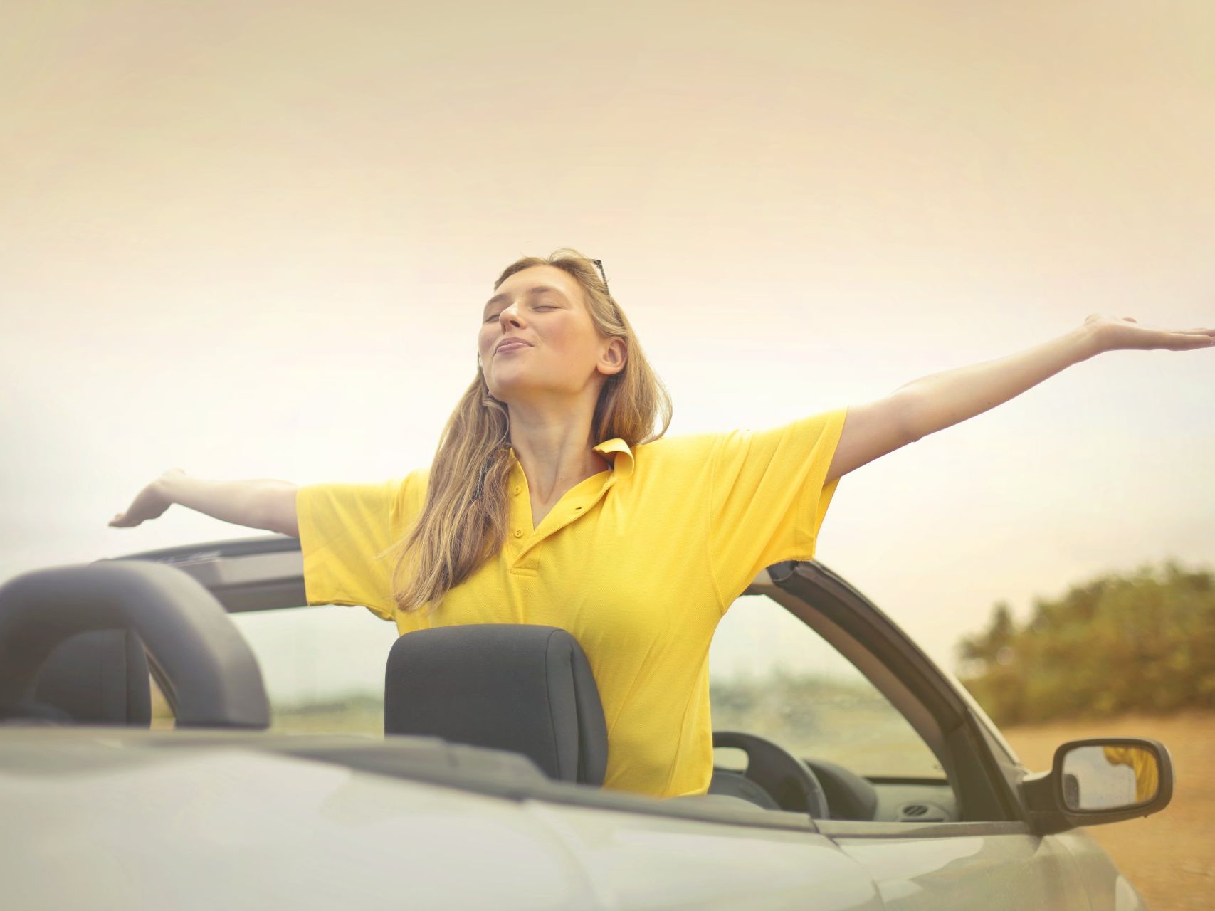 A woman in a yellow shirt is sitting in a convertible car with her arms outstretched.