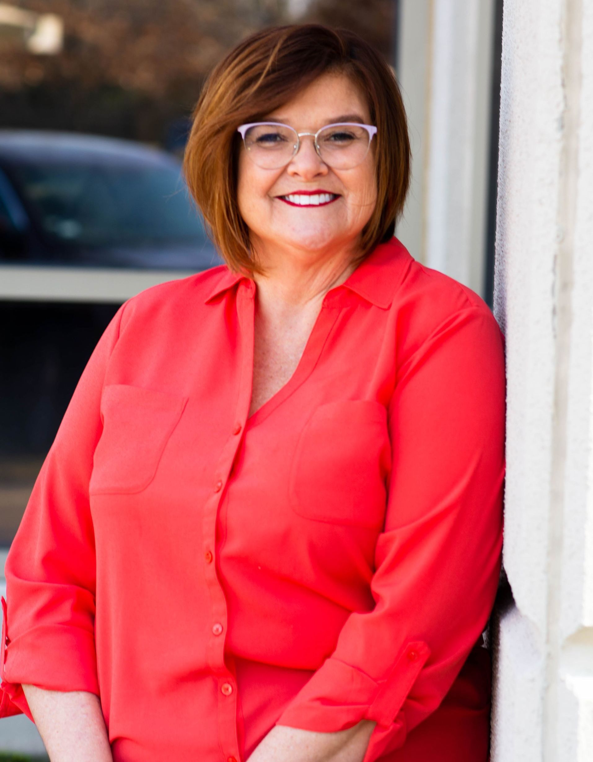 Karen Collom Picture A woman in a red shirt and glasses is leaning against a wall.