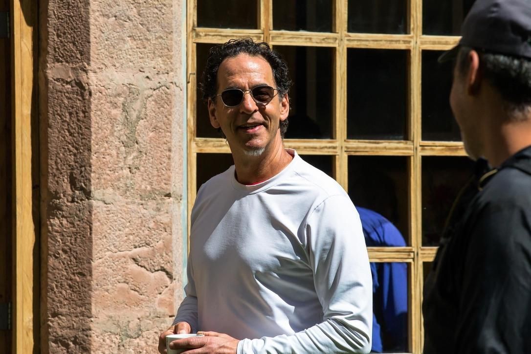 A man wearing sunglasses and a white shirt is standing in front of a building.