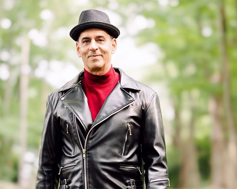 A man wearing a hat and a leather jacket is standing in the woods.