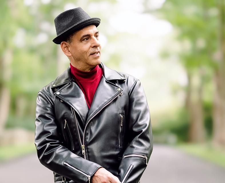 A man wearing a hat and a leather jacket is standing on a road.