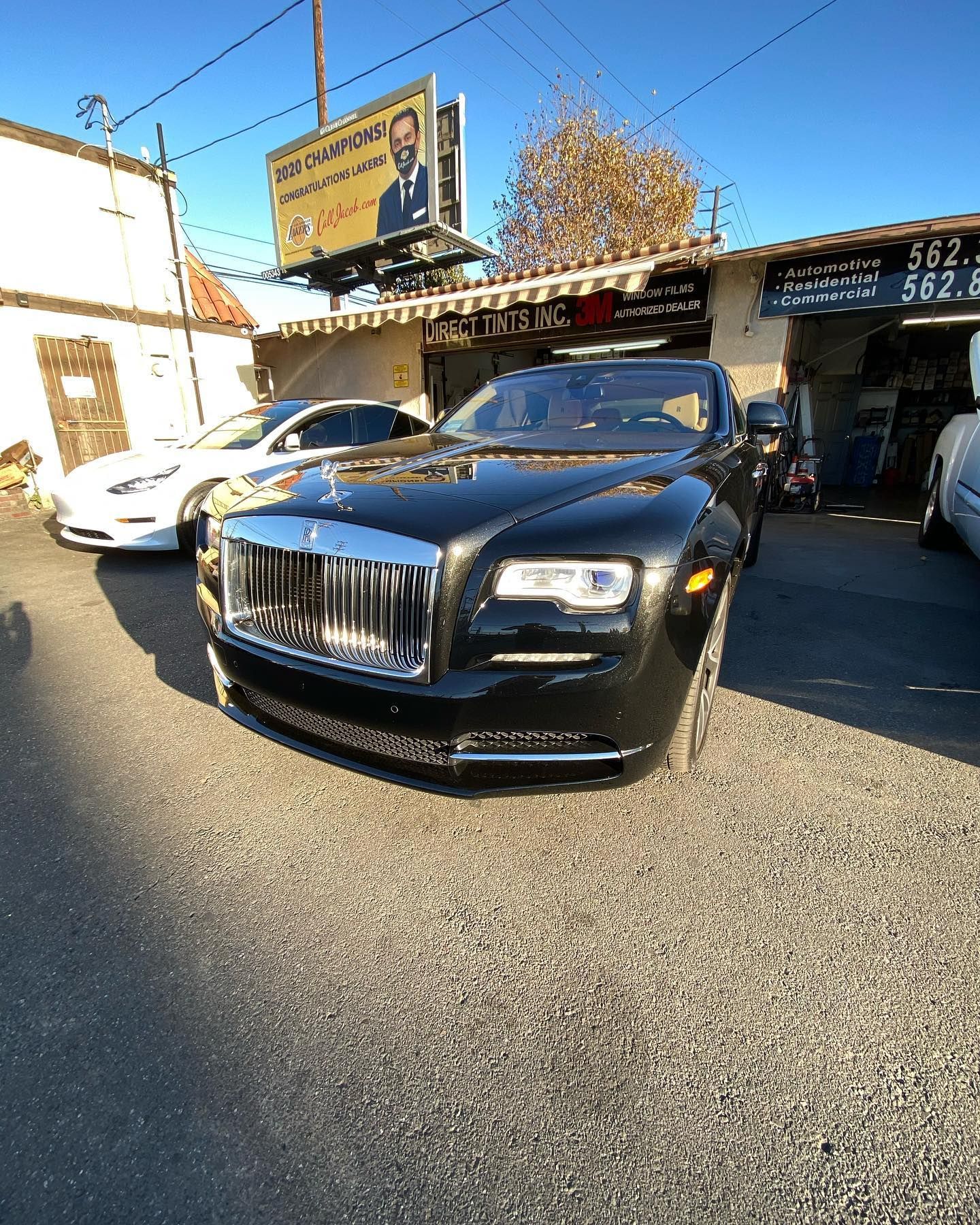 Black Rolls-Royce parked in front of a shop; white car behind. Sunny day.