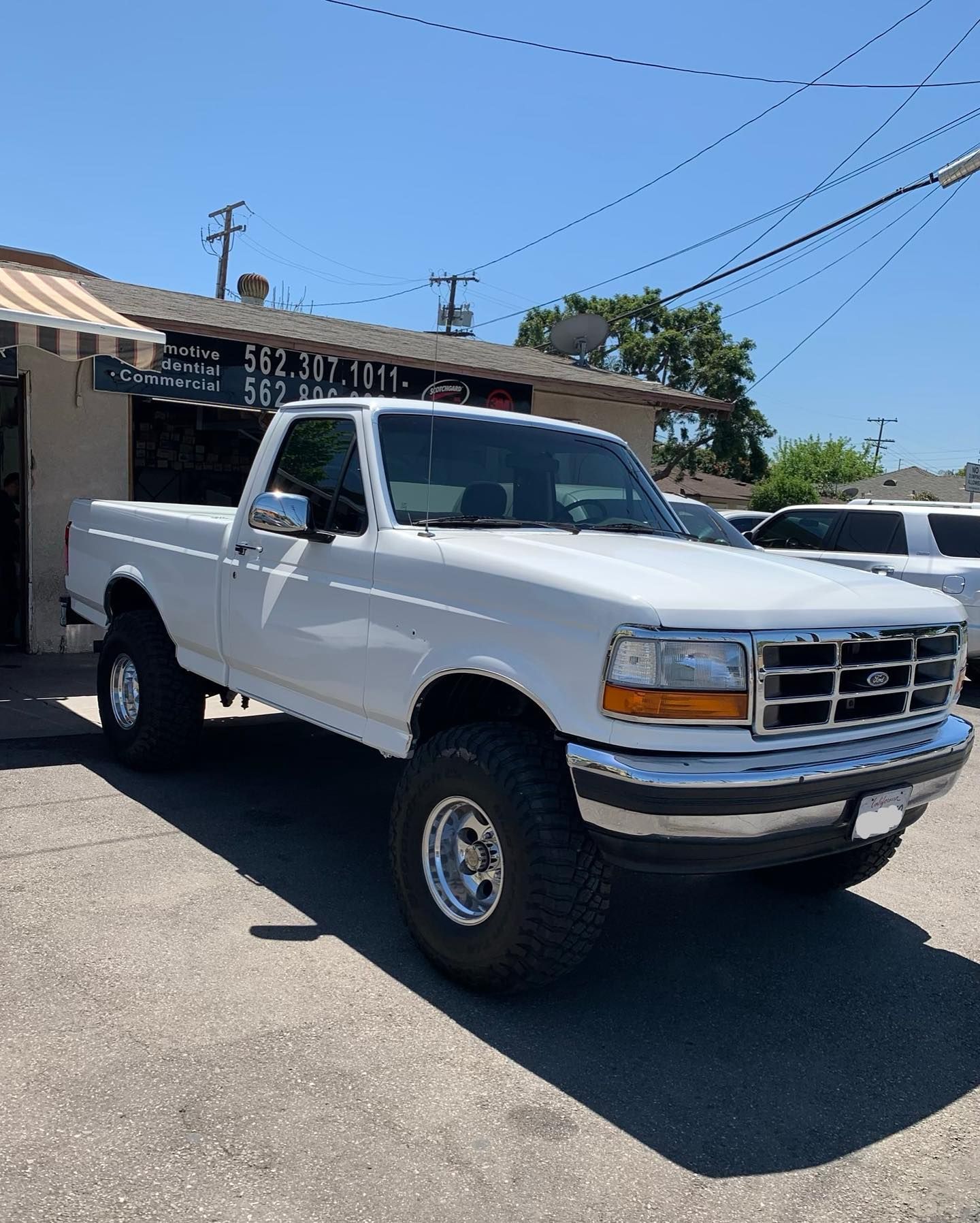 White Ford pickup truck with large tires parked in front of a building on a sunny day.