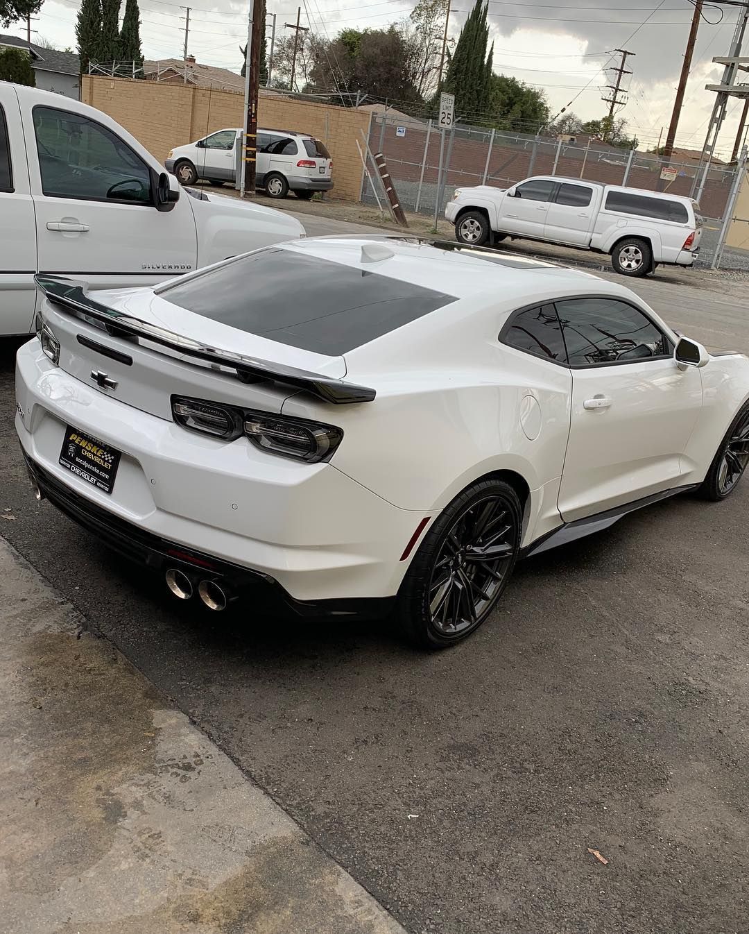 White Chevrolet Camaro parked on asphalt. Black wheels and spoiler. Other vehicles in background.