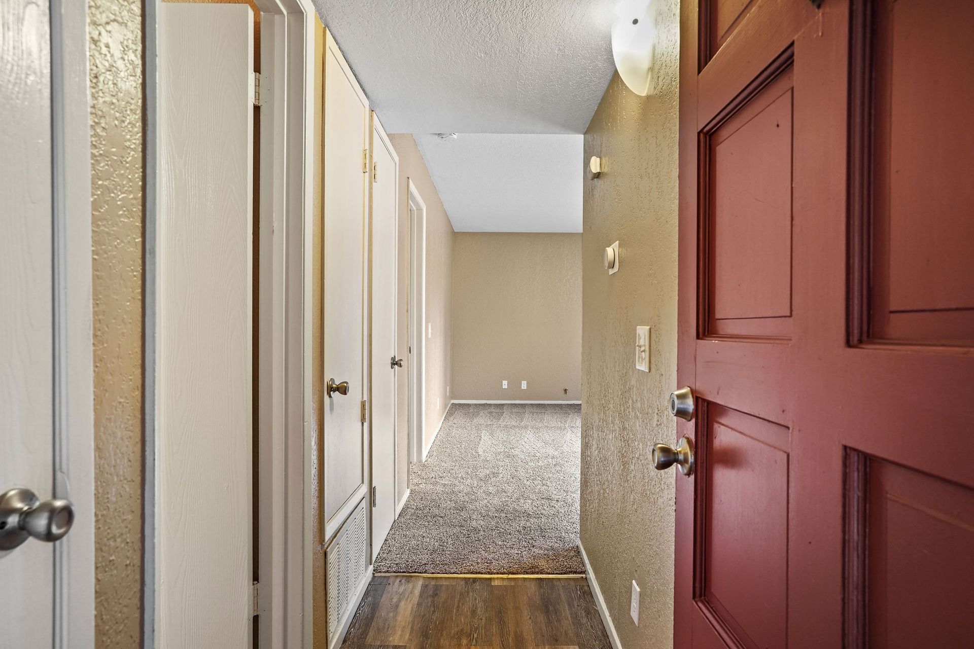 Narrow apartment hallway with white doors, beige walls, and a red front door in the foreground.